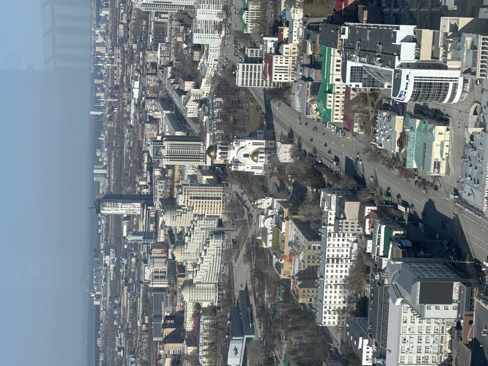 The image shows a high-angle view of Yekaterinburg, Russia, oriented vertically. The cityscape features a mix of high-rise buildings and low-rise structures, interspersed with trees and roadways. A prominent street runs through the center of the view, with cars visible on it. A large white building with golden domes is visible in the mid-ground, likely a cathedral. There are no people clearly identifiable in the image due to the distance. The environment appears to be urban, with a hazy sky above. The location is likely the city center of Yekaterinburg, Russia.
