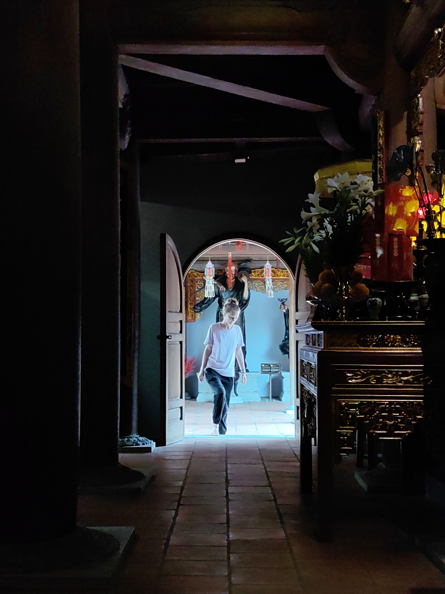 The image depicts a scene within a temple or religious structure in Hong Gai Ward, Vietnam. A young woman with her hair tied up is walking towards the viewer through an arched doorway. She is wearing a light-colored t-shirt and dark pants. Behind her, visible through the doorway, are two statues of figures in dark robes, likely deities or significant historical figures. Hanging between the statues are decorative lanterns or streamers. To the right of the doorway is a highly decorated wooden altar or table, adorned with flowers, fruits, and other religious objects, including a large red lantern with writing on it. The scene is framed by dark pillars and the interior of the building, suggesting a low-lit and possibly enclosed space. The floor is made of reddish-brown tiles, creating a sense of depth. The overall composition draws the eye towards the brightly lit doorway and the figure walking through it.