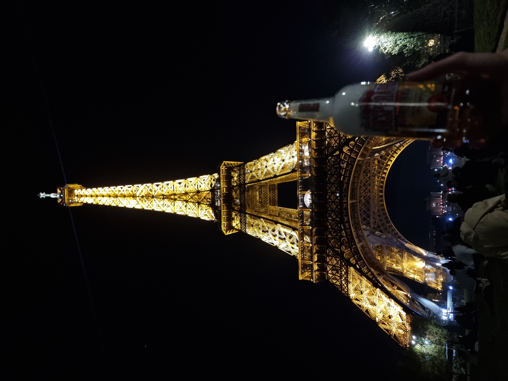 The image shows the Eiffel Tower in Paris, France at night. The tower is illuminated with yellow lights, and the sky is dark. A bottle of alcohol is being held in front of the tower. People are gathered near the base of the tower. The image is oriented vertically.