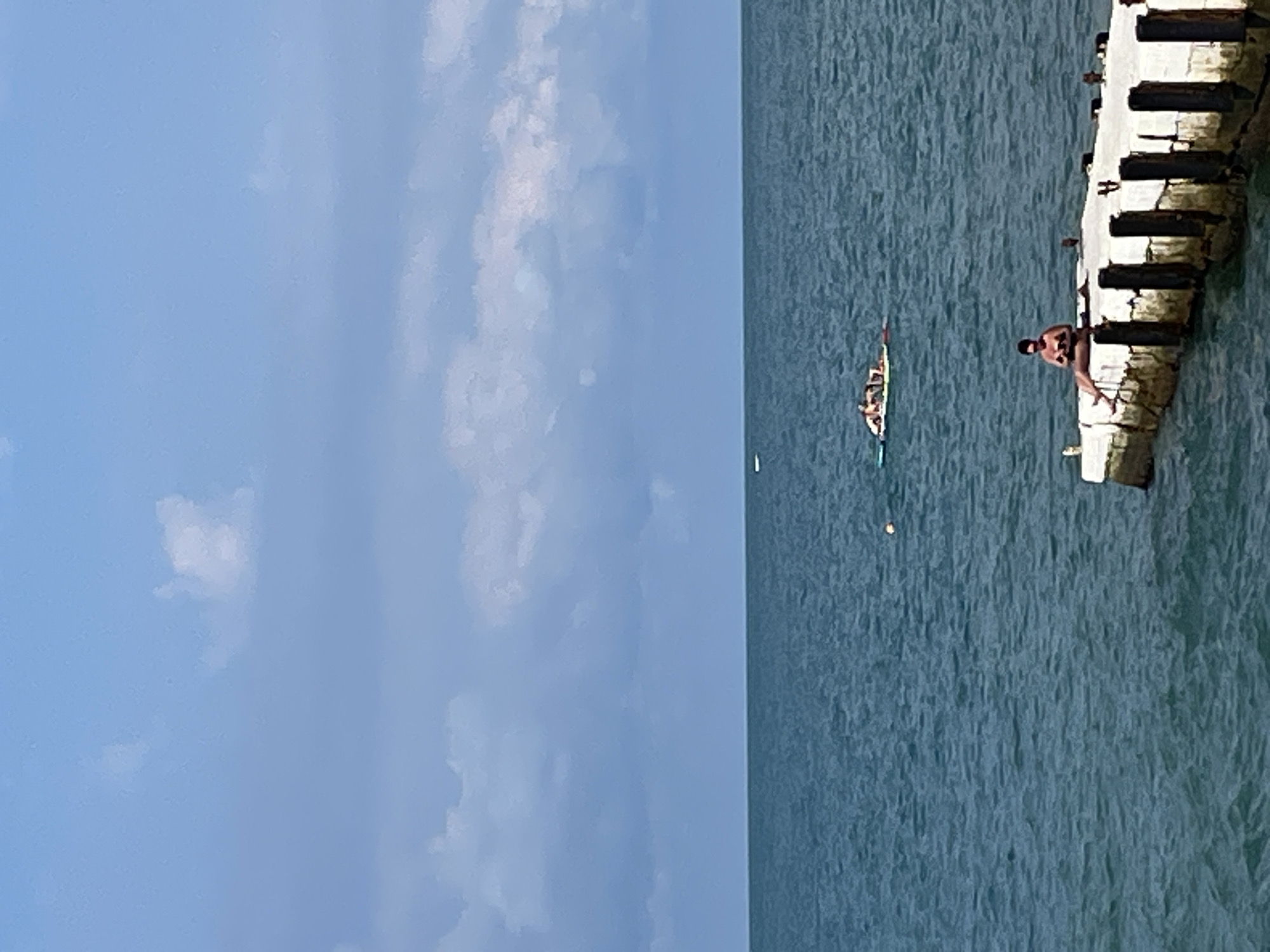 In Sochi, Russia, on a clear day with scattered clouds, a man is seated on the edge of a concrete pier, reading a book. The pier extends into the blue sea. Further out on the water, a group of people are relaxing on a paddleboard. The scene is serene and indicative of a typical summer day at the seaside.