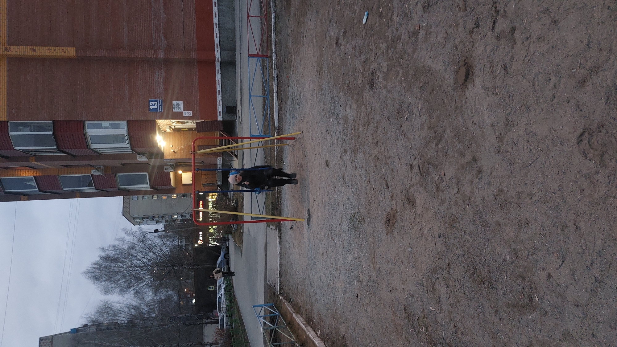 The image shows a playground in Novosibirsk, Russia, at dusk. A person is on a swing. The playground is adjacent to a brick apartment building. To the left of the playground, there is a view of a street with cars and trees. The ground is dirt. A building is labeled as number 13.