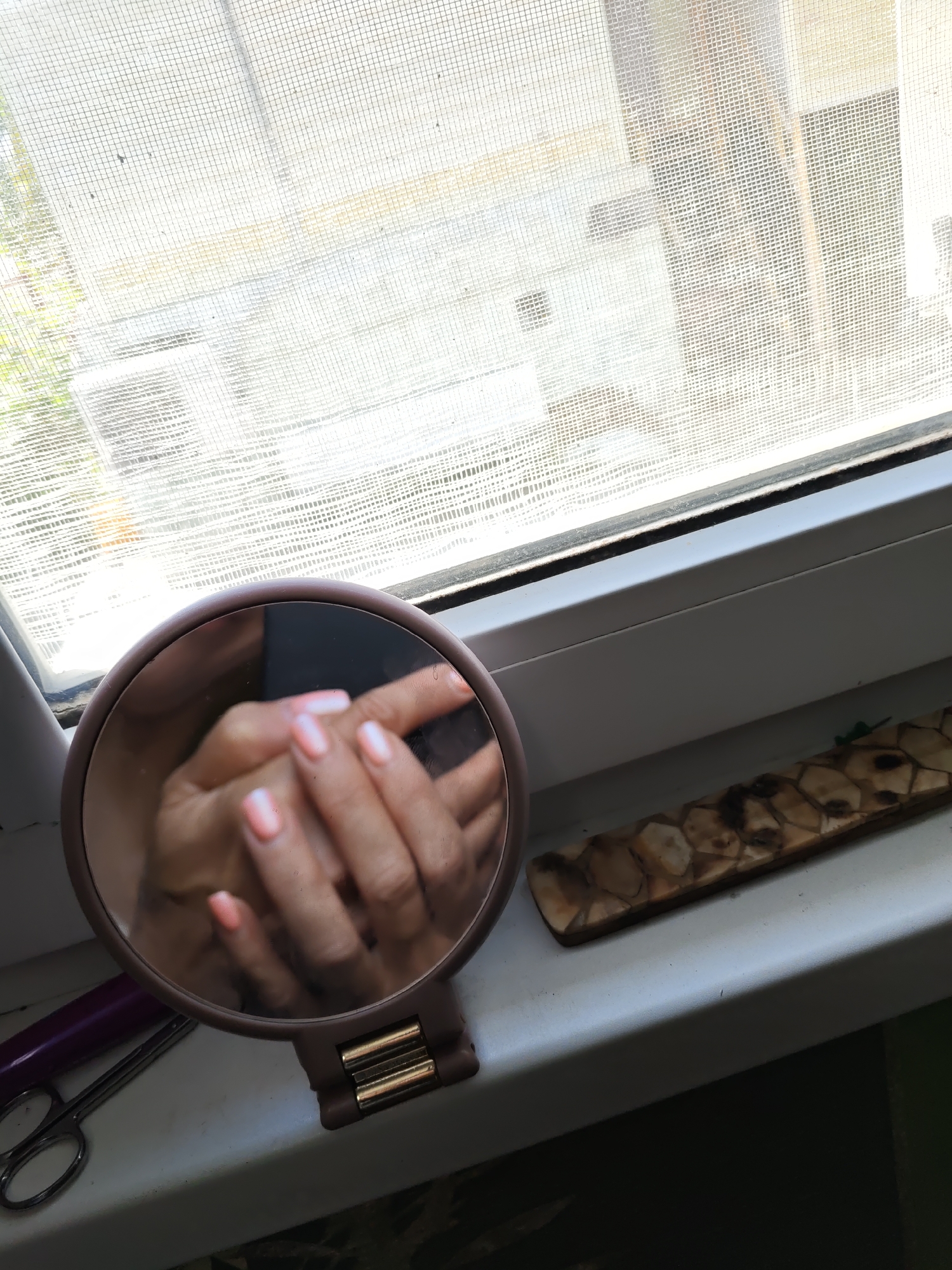 A close-up, high-angle shot shows a round mirror on a windowsill. The mirror reflects a pair of hands with light pink manicured nails, crossed over each other. To the left of the mirror are a pair of silver scissors and a purple object, possibly a brush handle. To the right of the mirror, a decorative wooden object with a mosaic-like pattern lies on the windowsill. The windowsill is illuminated by natural light coming through a window covered with a white mesh screen. Outside the window, hints of greenery and possibly buildings are visible, blurred by the screen. The scene appears to be an indoor setting, likely a room with a window overlooking an outdoor environment. The image was taken in Rostov-on-Don, Russia.
