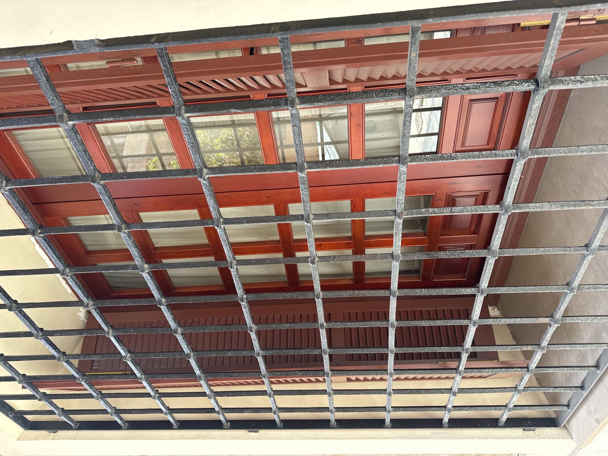 The image shows a close-up, angled view of a red wooden window with shutters behind a black metal security grate. The window has multiple panes and decorative wooden panels. The shutters are partially open, revealing slatted textures. Reflections are visible in the glass, including some greenery and a paved surface. The exterior wall of the building is a light cream color. The scene appears to be an outdoor view of a building in Murcia, Spain, likely on a street or courtyard. There are no people or animals visible in the image.