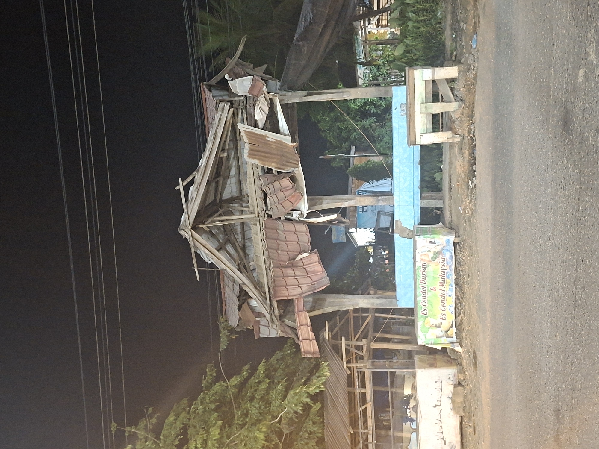 The image depicts a damaged outdoor structure in Keudeteupinpunti, Indonesia. The structure, possibly a food stall, has a partially collapsed roof made of corrugated metal and tiles. The wooden support beams are exposed and splintered. In front of the structure, a banner displays "Es Cendol Durian & Es Cendol Malaysia," indicating it was likely a place selling beverages. A low blue counter runs along the front, and a wooden stool or bench is positioned to the right. The ground in front of the stall is littered with debris. The background shows trees and other structures, partially obscured by darkness and the damaged stall. Power lines are visible overhead. The scene appears to be at night, with artificial lighting present. There are no people visible in the image.
