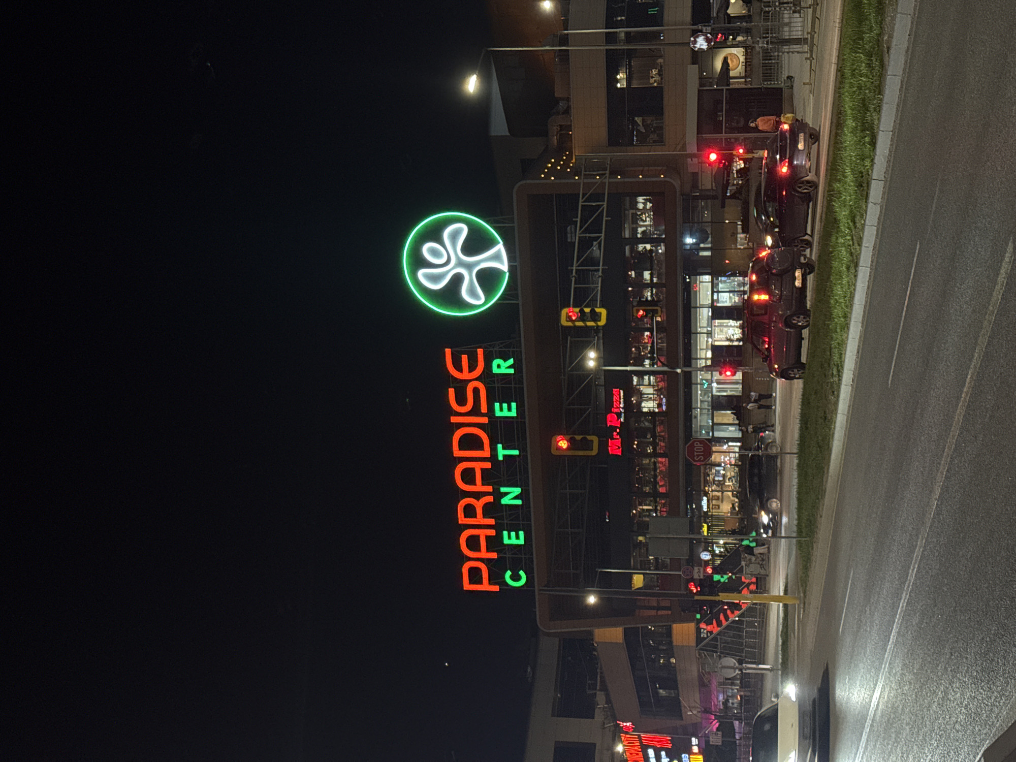 The image depicts Paradise Center, a shopping mall in Sofia, Bulgaria at night. The mall's illuminated sign is prominently displayed. Several cars are stopped at a traffic light on a road adjacent to the mall. There are streetlights and other building lights illuminating the area. People are visible in the background.