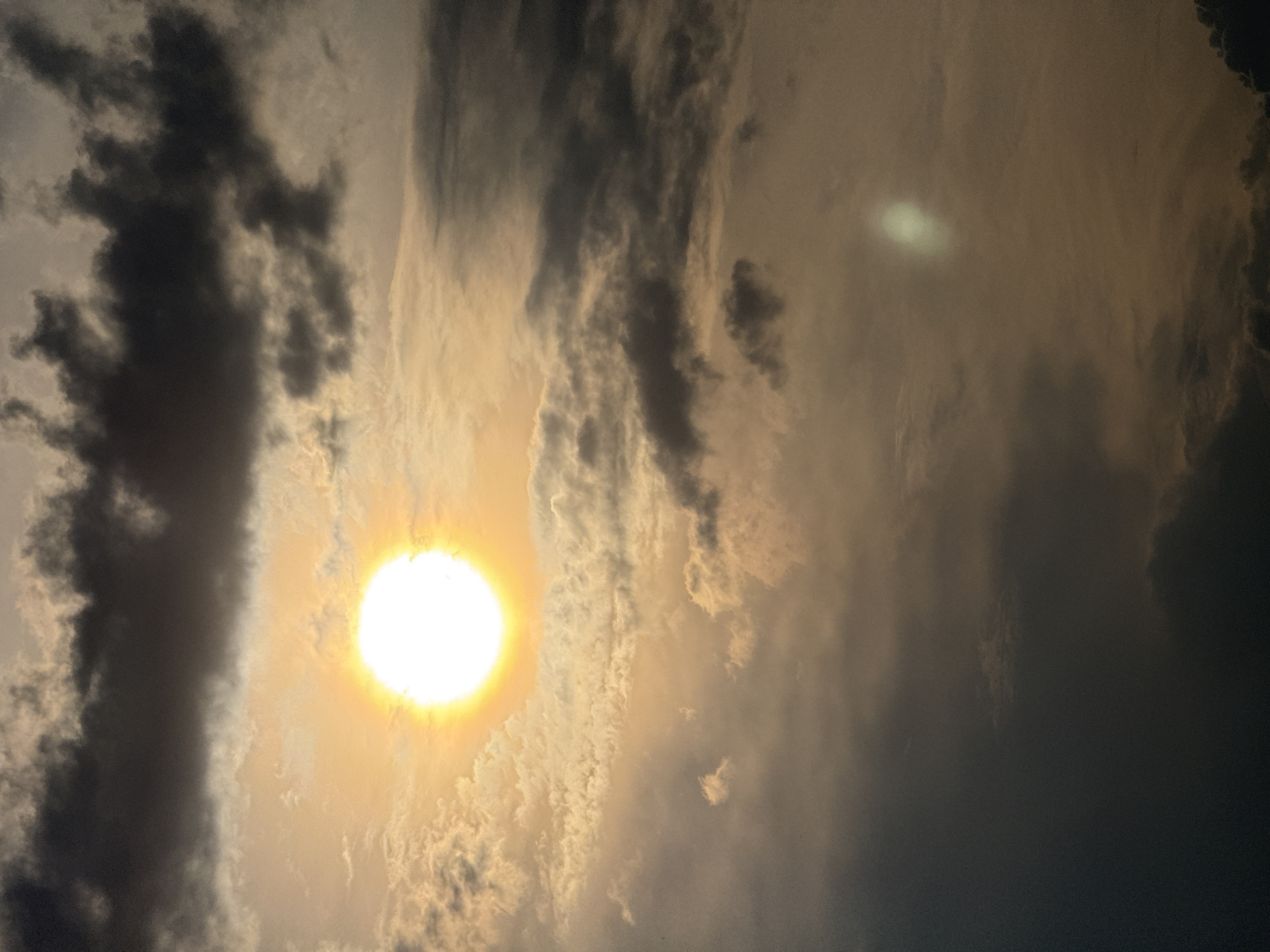 The image displays a cloudy sky with the sun prominently featured, appearing as a bright, glowing orb with rays of light emanating outwards. Dark, ominous clouds dominate the upper portion of the frame, while lighter, wispy clouds intermingle with the sun's glow. The overall lighting suggests either sunrise or sunset, casting a warm, golden hue across the scene. There are no discernible objects or people present in the image. The setting is an outdoor environment, likely a sky view. The provided context of Moscow, Russia, is not visually evident in the photograph itself.