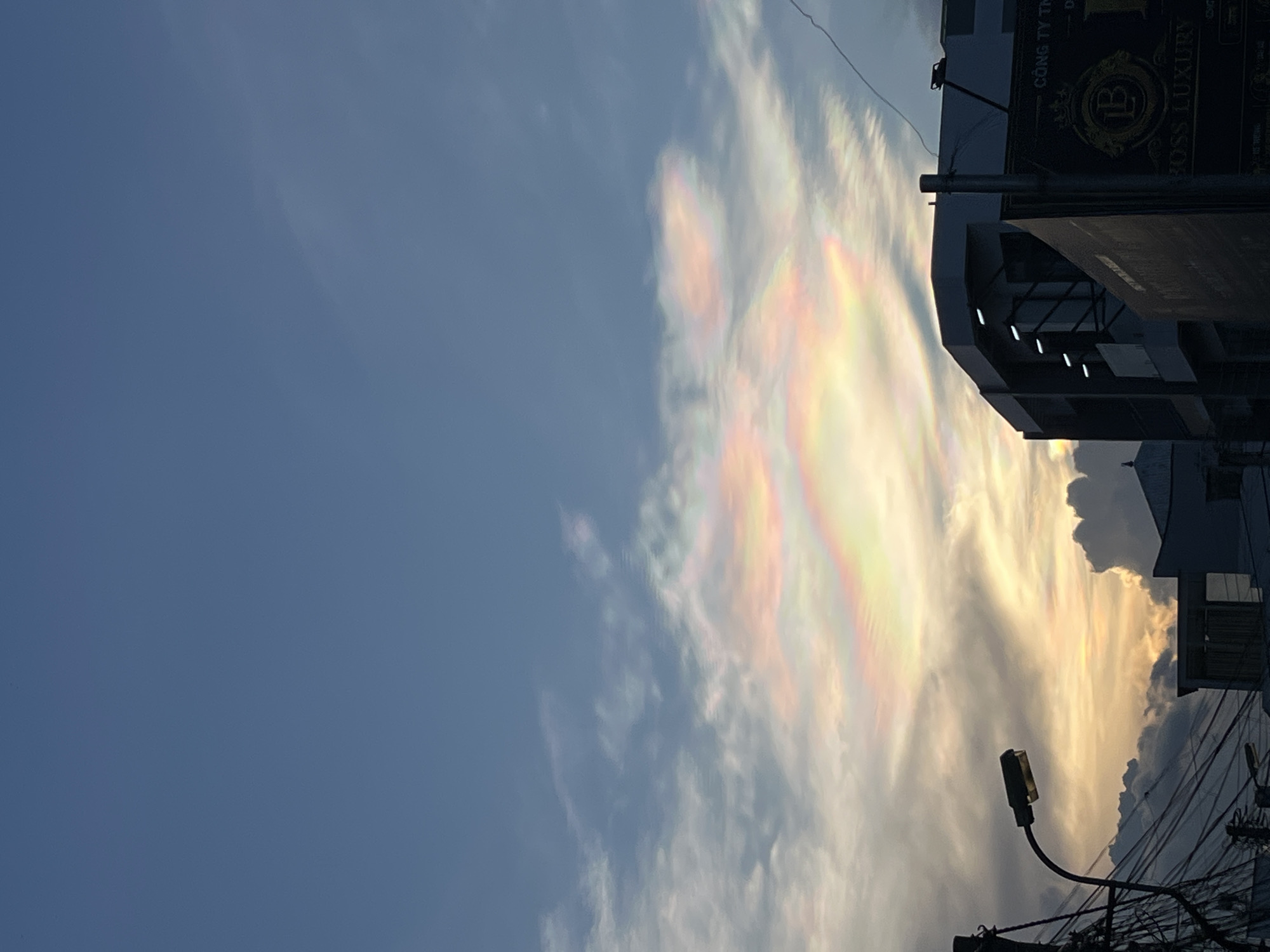 The image shows a view from Bac Nha Trang Ward, Vietnam, looking up at the sky, where a patch of iridescent clouds is visible. The sky is partially clear with a blue hue on the left side of the image. The iridescent clouds are on the right, displaying rainbow-like colors. At the lower right and partially obstructing the view of the sky, there's a streetlight. In the background on the right, there's a building with a sign that reads "BOSS LUXURY." Electrical wires are tangled in the lower right corner. The overall scene is captured during daytime, with the sun providing illumination to the clouds.