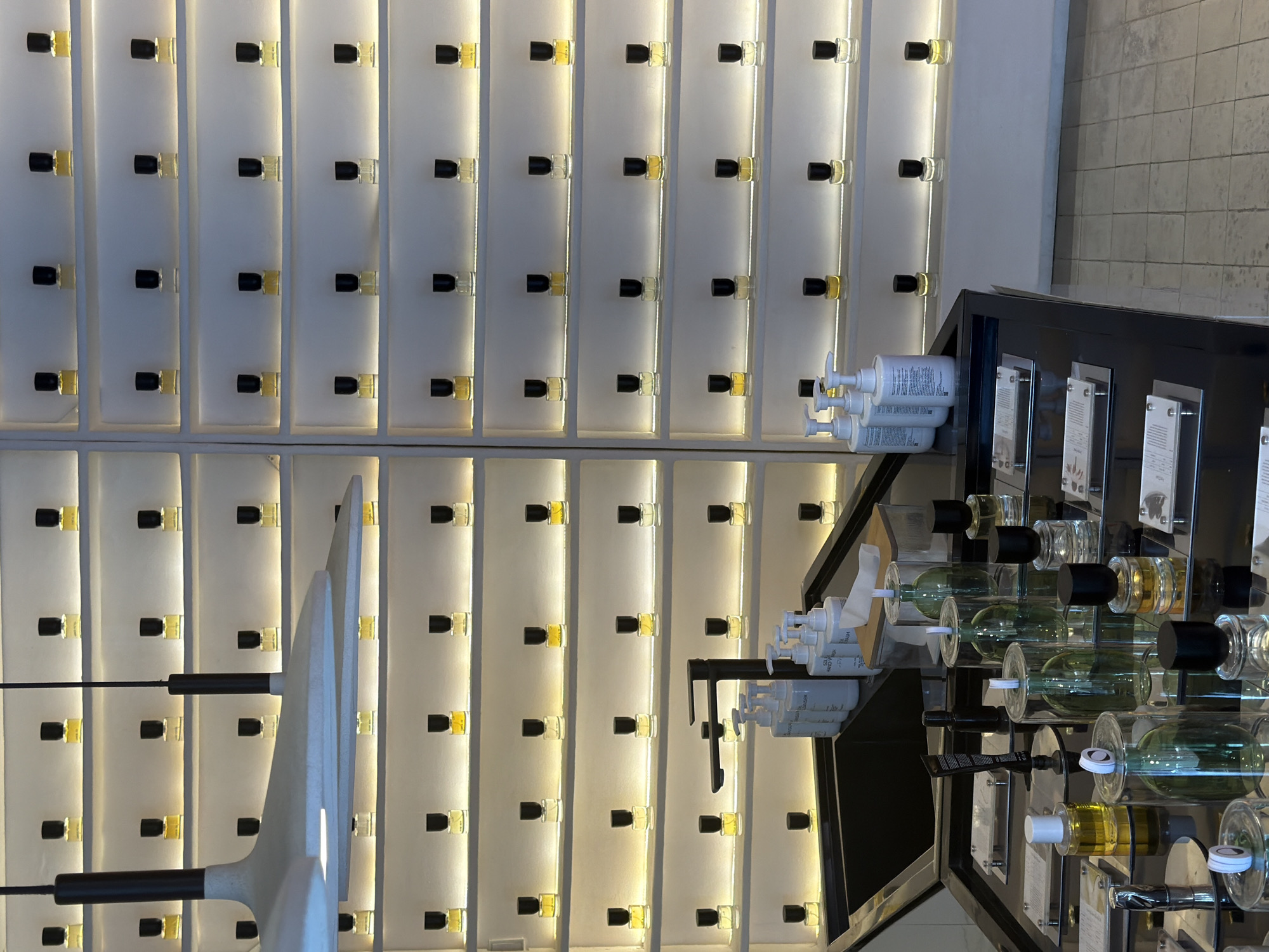 In Mengwi, Indonesia, a brightly lit store displays an array of perfume bottles on tiered shelving units. The shelves are illuminated from behind, casting a soft glow on the miniature bottles, most of which appear to contain yellow or clear liquids and are capped with black lids. The bottles are arranged in neat rows and columns.
In the foreground, a dark countertop holds various items. Several clear glass bottles, some containing green liquid and others clear, are prominent. Among these are several pumps dispensers, some labeled with white text. A black faucet with a modern design is positioned above a dark sink. On the countertop, there are also smaller perfume bottles, some with yellow lids and clear liquid, and others with black caps. Several informational cards are placed on stands in front of some of the display items. The overall environment suggests a retail setting focused on fragrance products. There are no people visible in the image.