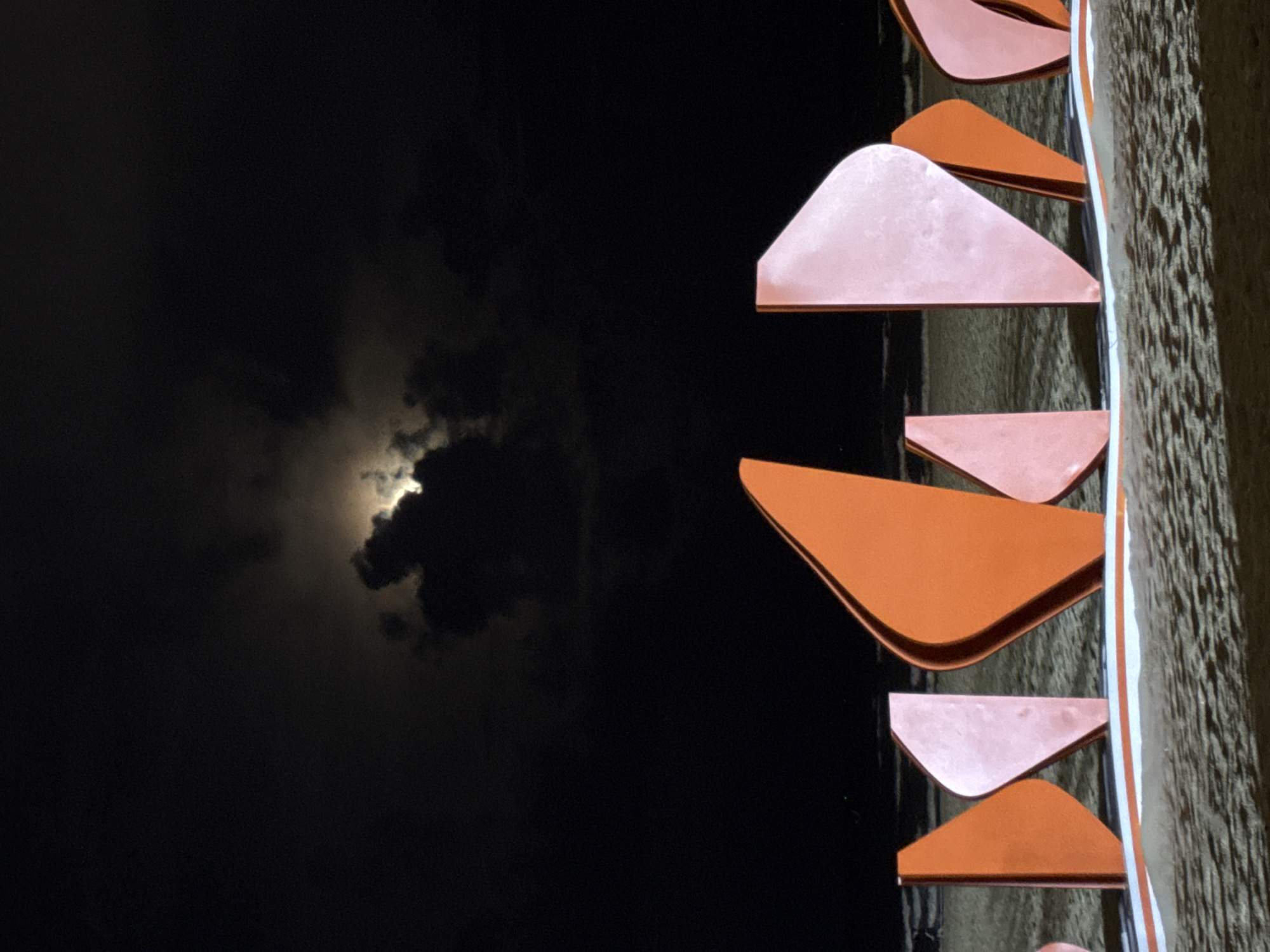 The image shows a nighttime scene on a beach in Da Nang, Vietnam. In the foreground, there is a row of abstract, colorful sculptures made of metal, alternating between orange and pink. These sculptures are positioned on the sand, with a white and orange striped base beneath them. Behind the sculptures, the ocean is visible, with gentle waves and the faint lights of distant structures or boats. The sky is dark, with a full moon partially obscured by dramatic, luminous clouds. The lighting suggests it is late at night, possibly illuminated by the moon and artificial lights from the sculptures or the city. There are no people visible in the frame.