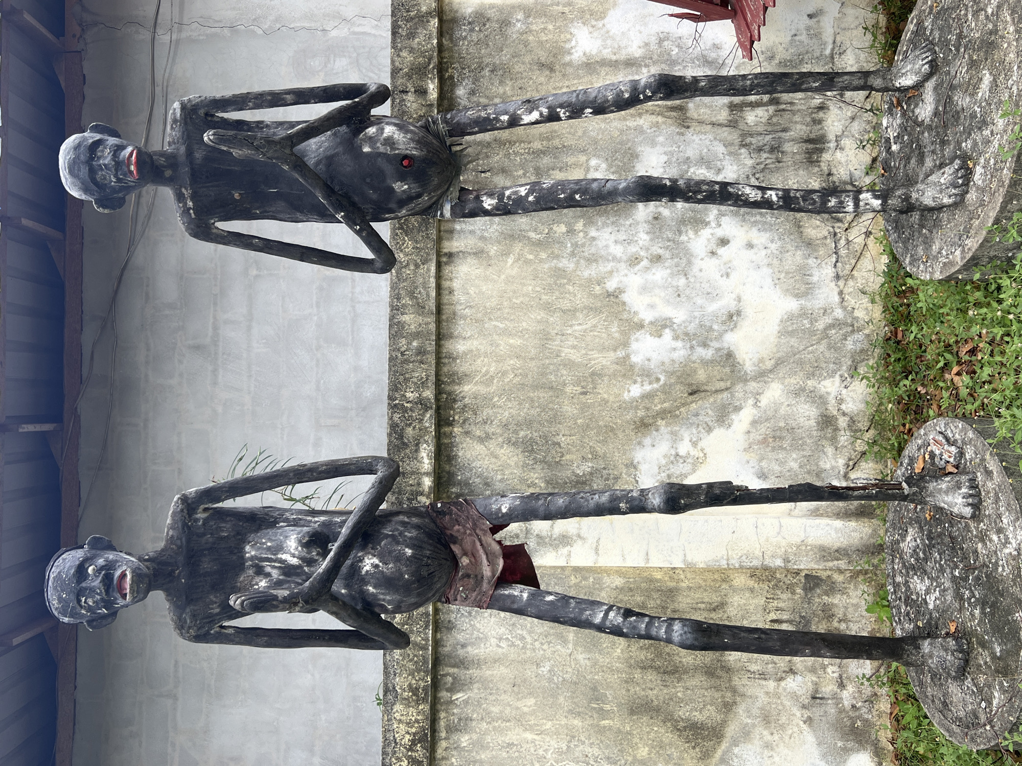 Two dark, emaciated statues with red lips stand on circular concrete pedestals in what appears to be an outdoor area. The statues are positioned side-by-side, facing the viewer, with their hands pressed together in a prayer-like gesture. They have large, disproportionate heads, slender limbs, and prominent bellies. The statue on the left wears a tattered, reddish cloth around its waist, while the statue on the right has a small red circular object embedded in its abdomen. Behind the statues is a weathered, light-colored wall with a textured surface. A corrugated metal roof is visible above the wall. The ground in front of the statues is covered with green grass and fallen leaves. The scene is located in Ko Pha-ngan District, Thailand.