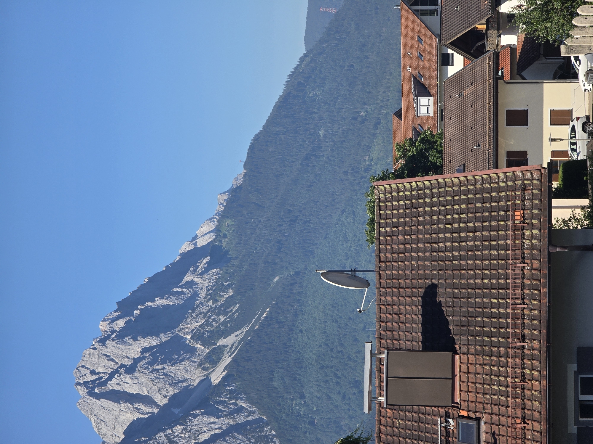 This is a telephoto shot taken from a residential area looking towards the Alps on a clear, sunny day. The foreground is dominated by the steeply pitched, tiled roofs of houses, with a satellite dish mounted on one of them. A dark, rectangular chimney or ventilation shaft is also visible on the closest roof. In the midground, more houses with red tile roofs and some trees are scattered. The background features a very large, imposing mountain range with jagged, rocky peaks. The lower slopes of the mountains are densely covered with dark green pine trees. The sky is a clear, bright blue, indicating good weather. The image appears to be taken in or near Garmisch-Partenkirchen, Germany, a town known for its proximity to the Bavarian Alps. No people or animals are visible in the image.
