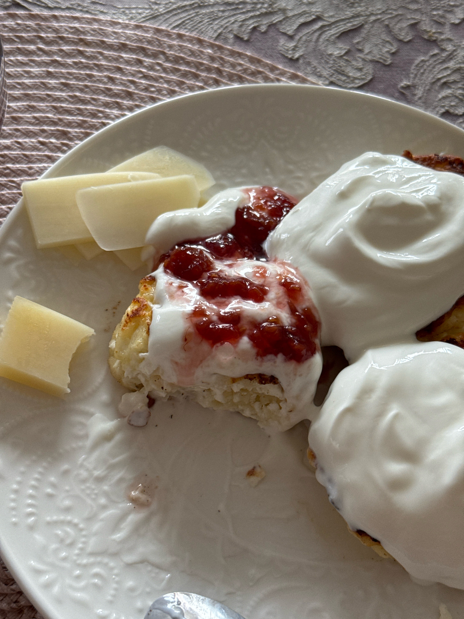 The image shows a close-up of a white plate with a textured surface, likely ceramic, featuring a meal. On the plate, there are several wedges of light yellow cheese. A small, round, golden-brown pastry or pancake is partially visible, topped with dollops of thick white cream or yogurt and a vibrant red jam, possibly strawberry or raspberry. More white cream is piled on the right side of the plate. The plate rests on a textured placemat with a horizontal striped pattern, which appears to be a light pink or beige color. The background shows a subtly patterned fabric, possibly a tablecloth, in a light purple hue. The lighting suggests an indoor setting, likely a home environment. There are no people visible in the image. The location in the city of Sinyavino, Russia, is not directly indicated by any visual cues in the image.