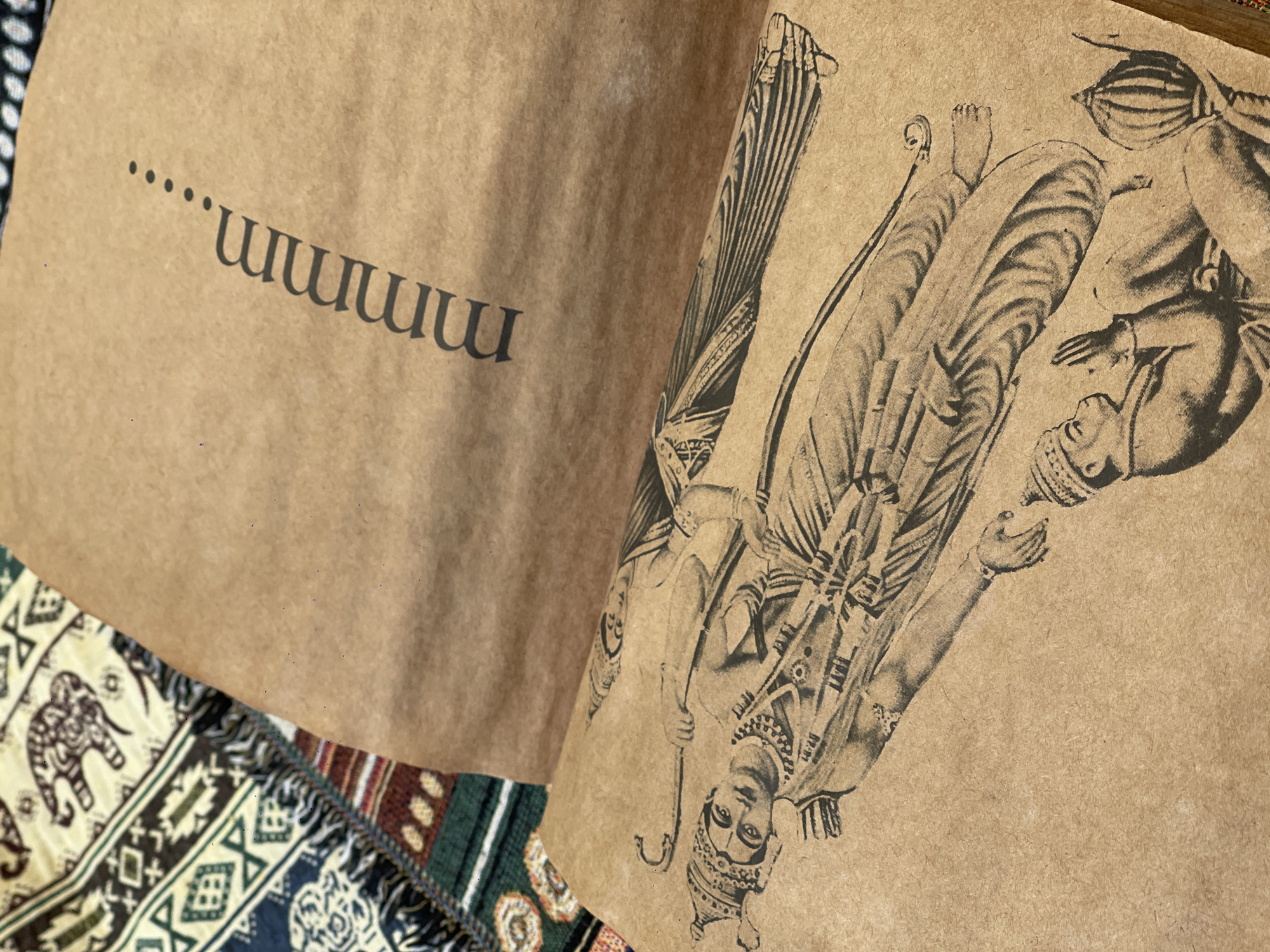 The image shows an open book with pages made of brown paper. The left page features a detailed black ink drawing of a reclining figure wearing ornate headwear and jewelry, holding a bow and arrow. Below this figure is a smaller drawing of a person in a seated position with hands clasped. The right page has the word "Mmmm" followed by ellipses printed in a darker shade of ink. The background consists of a colorful, woven textile with geometric patterns and depictions of elephants. The image does not contain any information about the city of Đà Nẵng, Vietnam.