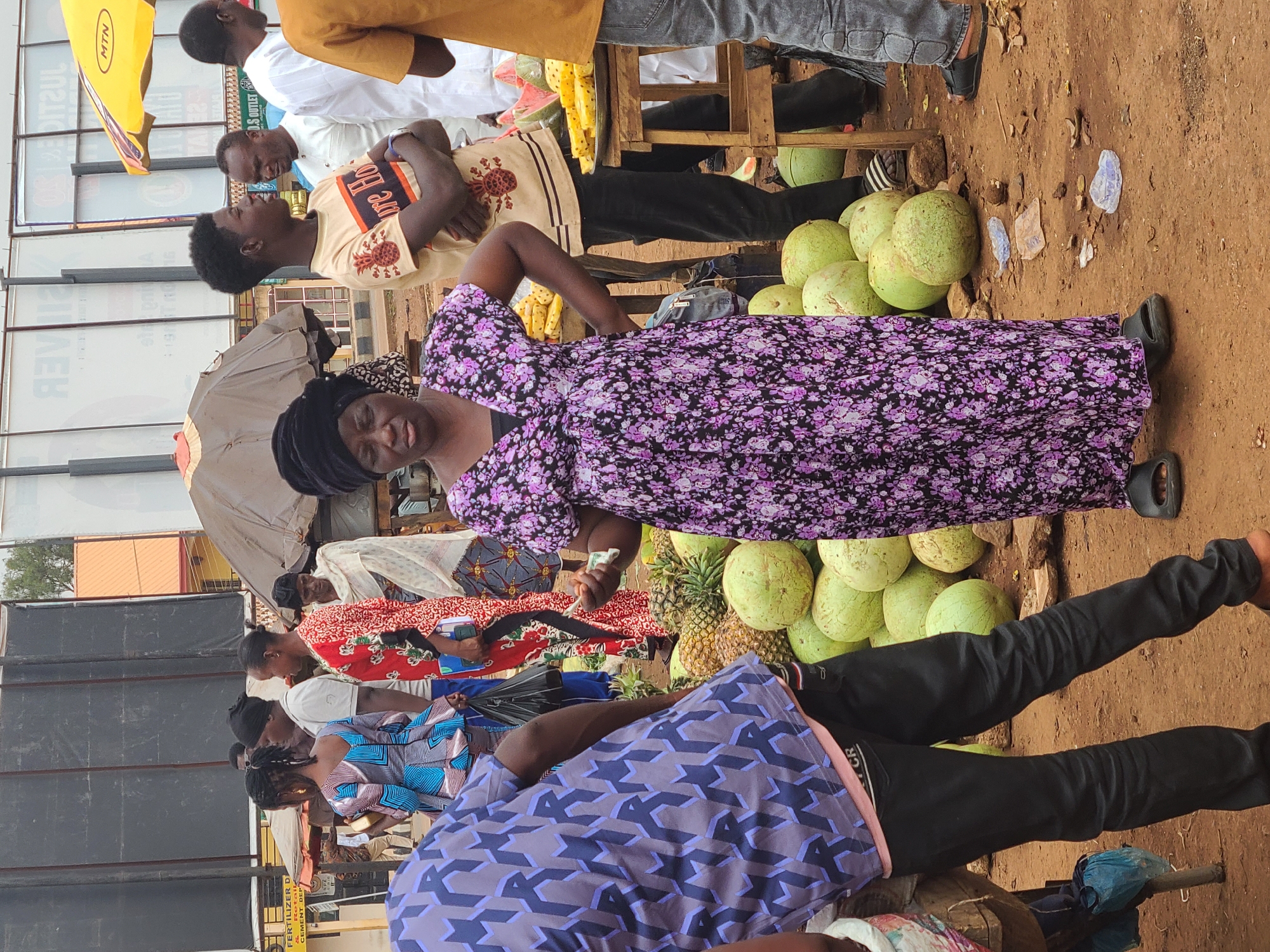 The image shows a bustling outdoor market scene in Jos, Nigeria. People are milling around, buying and selling goods. A woman in a long, purple floral dress stands prominently, holding a piece of currency in her hand. Stacks of green fruits, likely gourds, are beside her, as well as pineapples. Other people are visible around her, some inspecting goods, others seemingly in conversation. Clothing styles vary. The market appears to be on a dirt ground. The environment also includes buildings in the background with commercial signage.