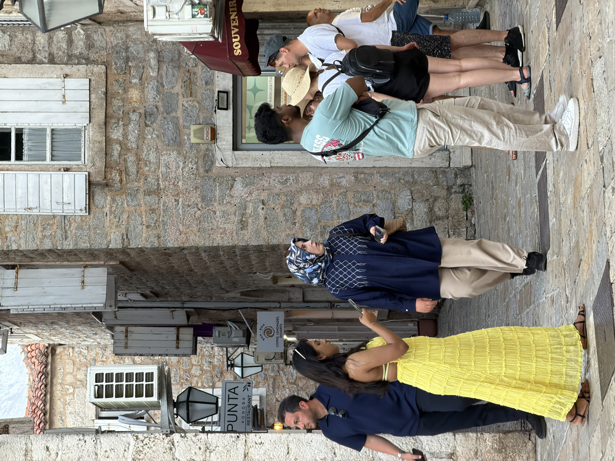 The image depicts a narrow, cobblestone street in the Old Town of Budva, Montenegro. The architecture features weathered stone buildings with wooden shutters, characteristic of the Mediterranean style. In the foreground, a group of people are walking and standing.
On the left, a man wearing a dark blue polo shirt and black trousers is walking away from the camera, holding a phone in his left hand. Beside him, a woman in a vibrant yellow dress is looking at her phone.
In the center, a woman wearing a dark blue abaya and a patterned hijab is walking, looking to her left. She is carrying a brown bag.
To the right, a man in a light blue t-shirt and beige trousers is standing with his arms crossed, facing towards the right. He has a black bag slung across his shoulder. Next to him, a woman in a straw hat and a white top is looking towards him. Behind them, another man in a white t-shirt and a baseball cap is also present, and further to the right, another man in a white t-shirt and patterned shorts is visible.
Signs for businesses are visible on the buildings. One sign reads "PUNTA KONOBA RESTAURANT," and another smaller sign indicates "BON-BON COFFEE, LEMONADES, DESSERTS." A red awning over a shop reads "SOUVENIR." An air conditioning unit is mounted on one of the buildings. The overall scene captures a moment of daily life on a busy street in the historic center of Budva.