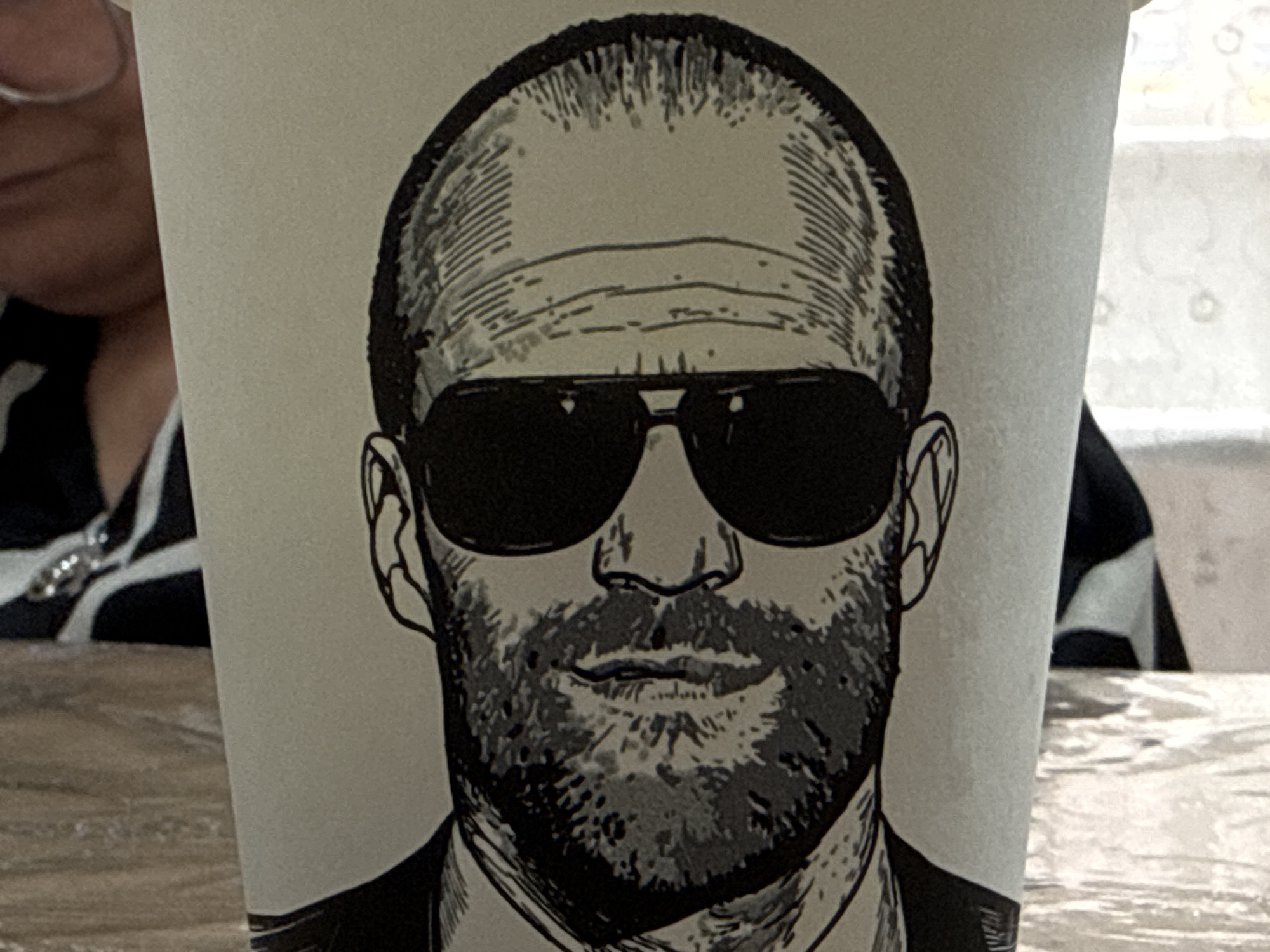 A black and white drawing of a man's face with sunglasses and a beard is printed on a paper coffee cup. The man's head is bald and his eyes are hidden behind the dark lenses of his sunglasses. He is wearing a suit jacket and a tie. The cup is placed on a wooden table. In the background, a person wearing glasses and a black and white striped shirt is visible. The scene is located on a table in the city of Beslan, Russia.