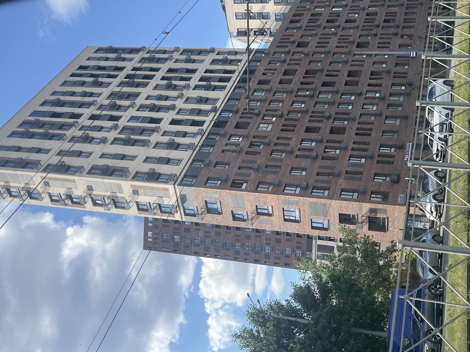 The image captures a view of modern, high-rise apartment buildings in Kazan, Russia. The primary building in the foreground is constructed with a combination of light-colored brick on its upper floors and reddish-brown brick on the lower sections. It features numerous windows, many with dark frames and some with balconies or recessed sections. A second, taller building with a similar brick pattern is visible behind and to the left, partially obscured by the foreground structure. The sky above is blue with scattered white clouds. In the foreground, there is a street with several cars, including a dark gray Audi SUV, a silver compact car, and a white hatchback. A blue van is also visible. A metal fence with a diamond pattern runs across the bottom of the image, with green grass and some foliage behind it. Power lines and utility poles are also present in the scene.