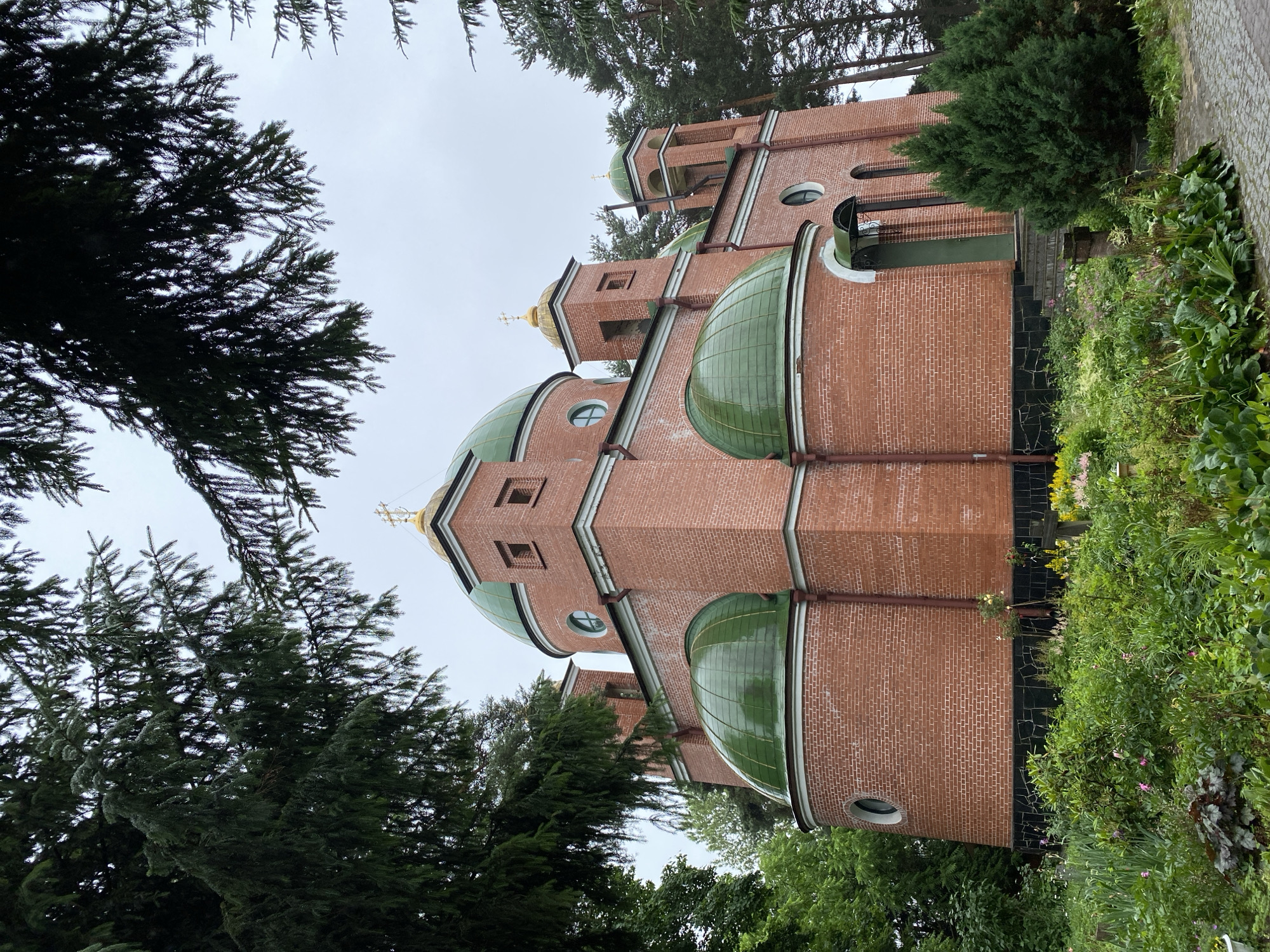 A brick church with green domes is situated in a densely wooded area, likely a cemetery, in Priozersk, Russia. The church is a prominent structure made of red brick, featuring multiple domes, some larger and some smaller, all covered in a reflective green material. Two bell towers rise from the sides of the main building, also topped with green domes and golden crosses. The church has arched windows and a main entrance with a green awning.
The foreground is filled with lush greenery and flowering plants, suggesting a well-maintained garden. The plants are varied in color and height, with some taller trees and bushes surrounding the church. In the background, tall pine trees frame the scene, indicating a natural, perhaps rural or park-like setting. The sky is overcast, contributing to a subdued lighting condition. There are no people visible in the image. The ground in front of the church appears to be paved with cobblestones, and a wooden cross is visible among the plants. The overall impression is one of a peaceful and somewhat secluded religious site within a natural environment.