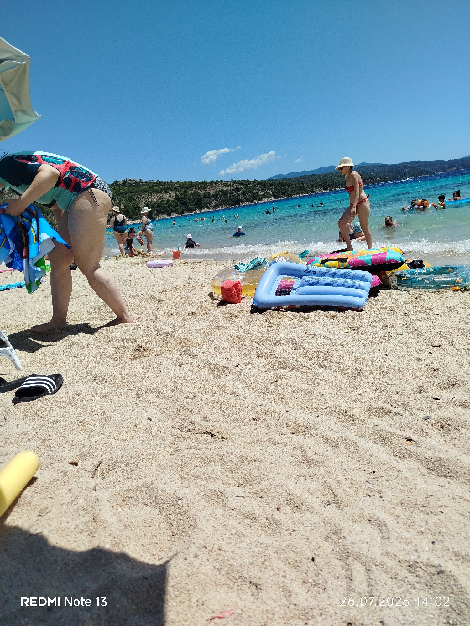 The image depicts a sandy beach on a sunny day, with people enjoying the water and beach activities. In the foreground, a person wearing a swimsuit is bending down, holding a towel. Several inflatable toys, including a blue raft and a smaller ring float, are scattered on the sand. In the background, the clear blue water is filled with swimmers, and a verdant coastline with trees and hills forms the backdrop. The sky is bright blue with a few scattered clouds. The overall scene suggests a typical summer day at a Greek beach, possibly within the Sithonia Municipal Unit, known for its beautiful coastal landscapes. The presence of beach umbrellas and the relaxed atmosphere further reinforce this setting.