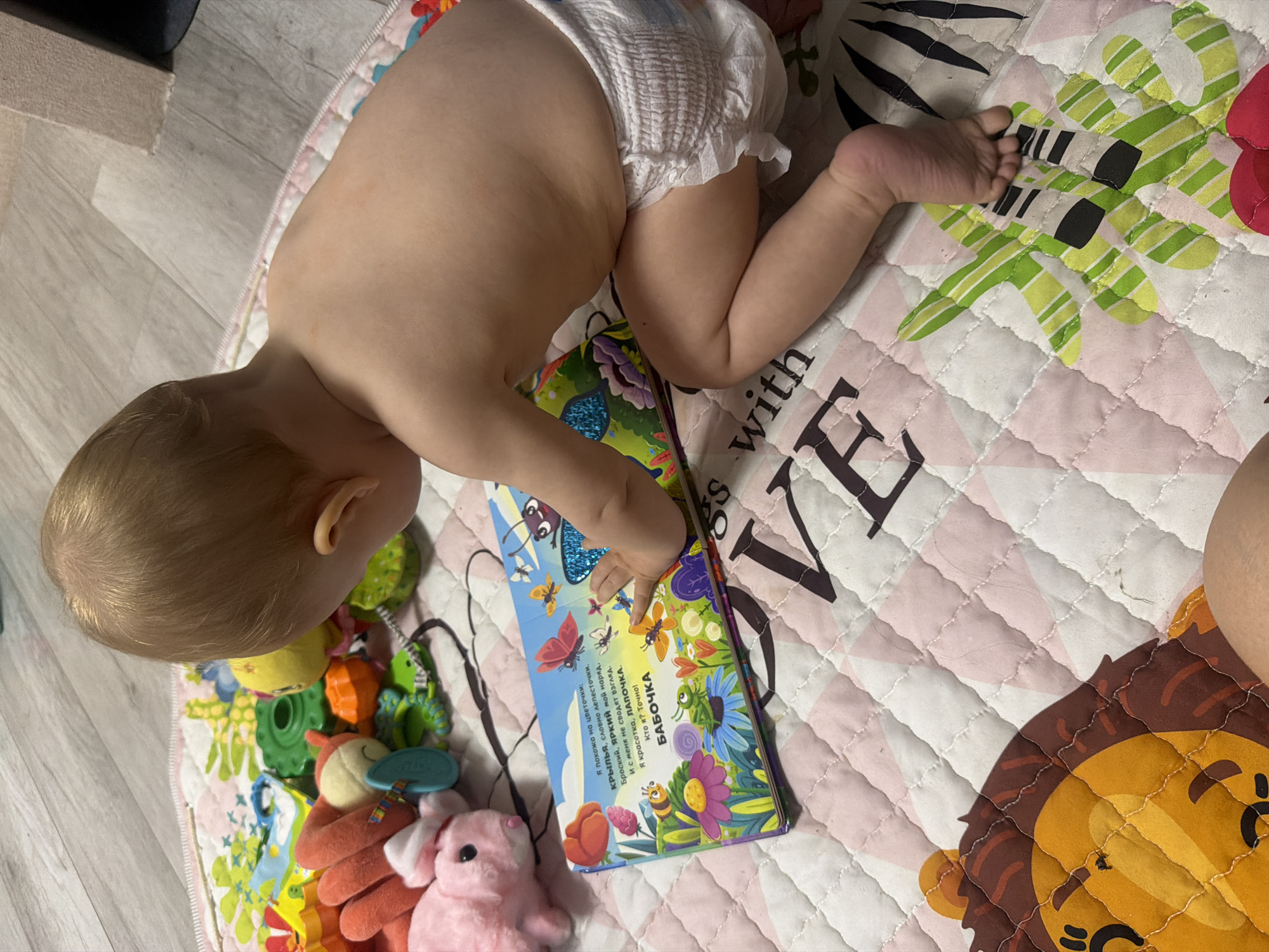 A baby is lying on a playmat, looking at a book. The baby is wearing a diaper and has blonde hair. The playmat is colorful and has animal prints on it. There are also several toys scattered around the baby. The book is open and the baby's hand is pointing at a page with a picture of a butterfly and text. The text is in Russian and appears to be a children's book. The overall scene is peaceful and suggests a moment of quiet play or learning for the baby. The lighting indicates it is daytime, and the indoor setting implies a home environment. The presence of a playmat and toys suggests this is a nursery or a child's play area. The text in Russian translates to: "I look like flowers: Wings, like petals. My outfit is bright, my bright. And they don't take their eyes off me. I am a beauty, a sweetheart. Who am I? Exactly! BUTTERFLY."