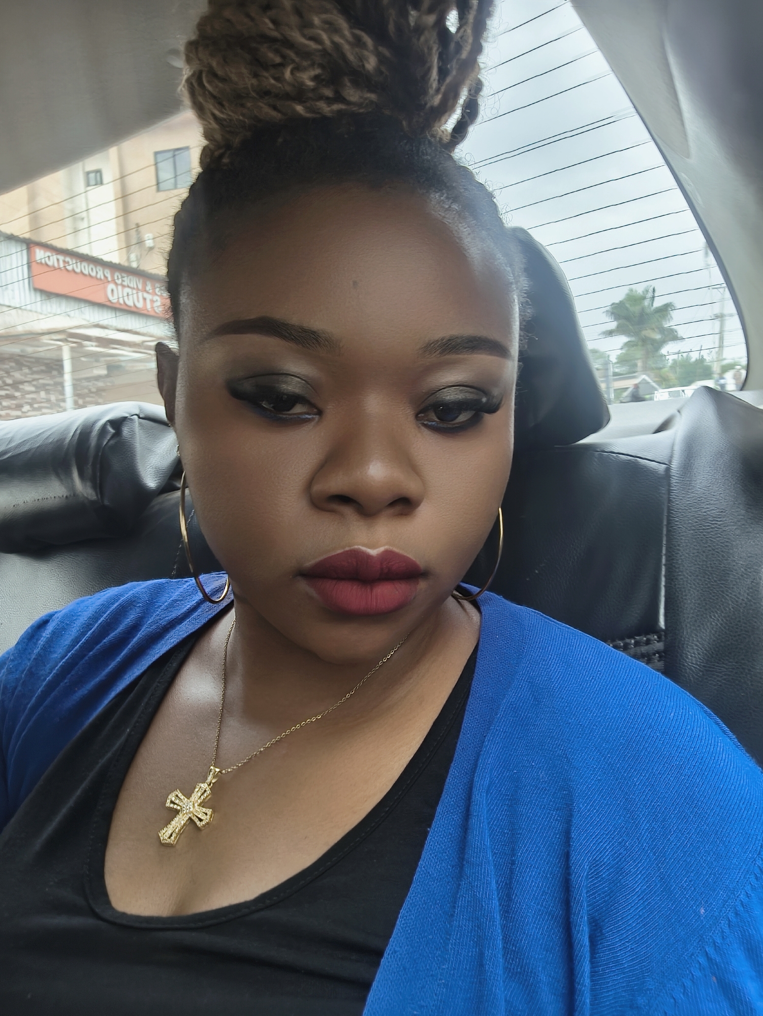 A woman is in the back seat of a car in Jos, Nigeria. The woman has her hair up in a bun and is wearing a gold cross necklace, gold hoop earrings, a black shirt, and a bright blue sweater. The sign for "Studio 2 Video Production" is visible through the window. A palm tree is also visible through the window.