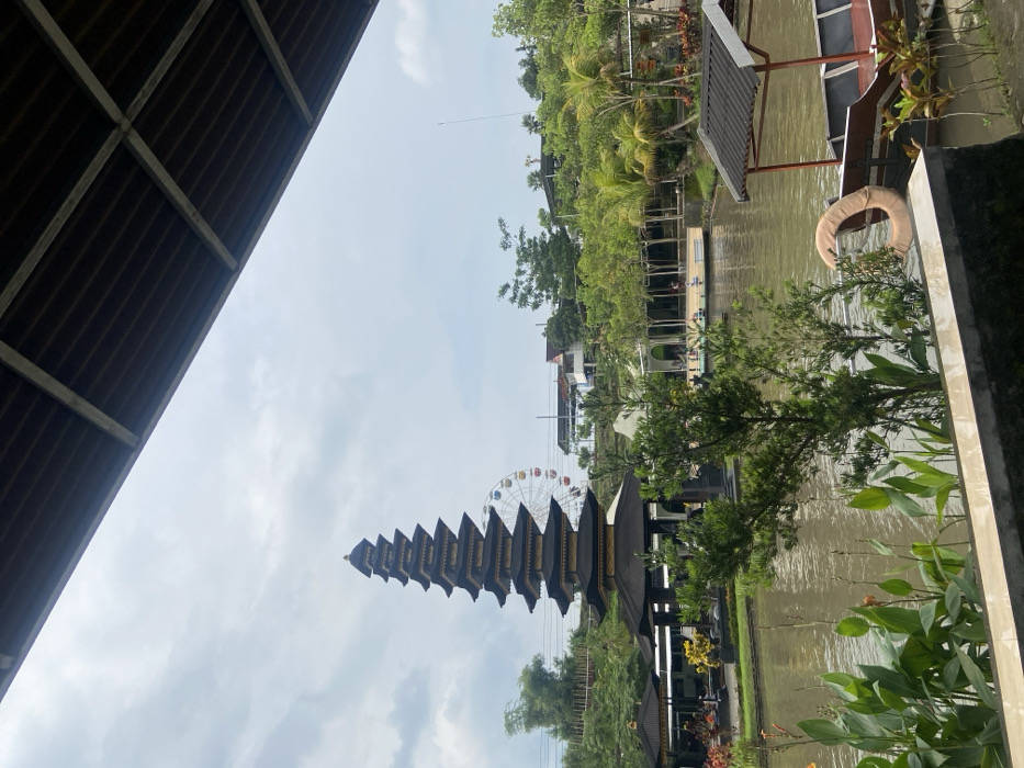 A tall, stepped pagoda-like structure with a Ferris wheel behind it stands against a cloudy sky. Lush green vegetation borders a body of water with small buildings and structures lining its edge. A boat with a canopy, a life preserver, and plants are visible. Part of a dark roof structure looms in the foreground. The scene appears to be in a park or tourist attraction.
