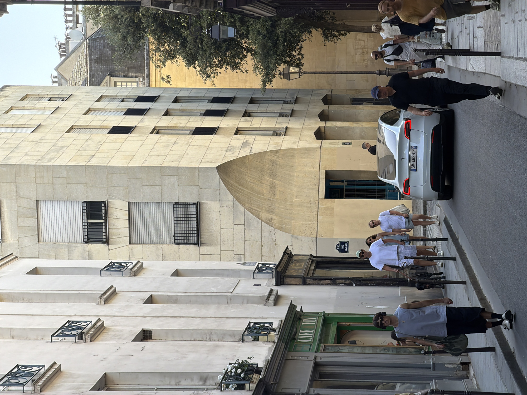 The image captures a street scene in Paris, France. A white Tesla is parked on the right side of a narrow street. A man in a black shirt and blue cap stands near the car. Several pedestrians are walking along the sidewalk. The buildings lining the street are old, with architectural details like wrought-iron window decorations and stone facades. One building features a curved arch. A sign indicating "Rue Elzevir" is visible on the side of one of the buildings. A cafe with a green awning is also present.