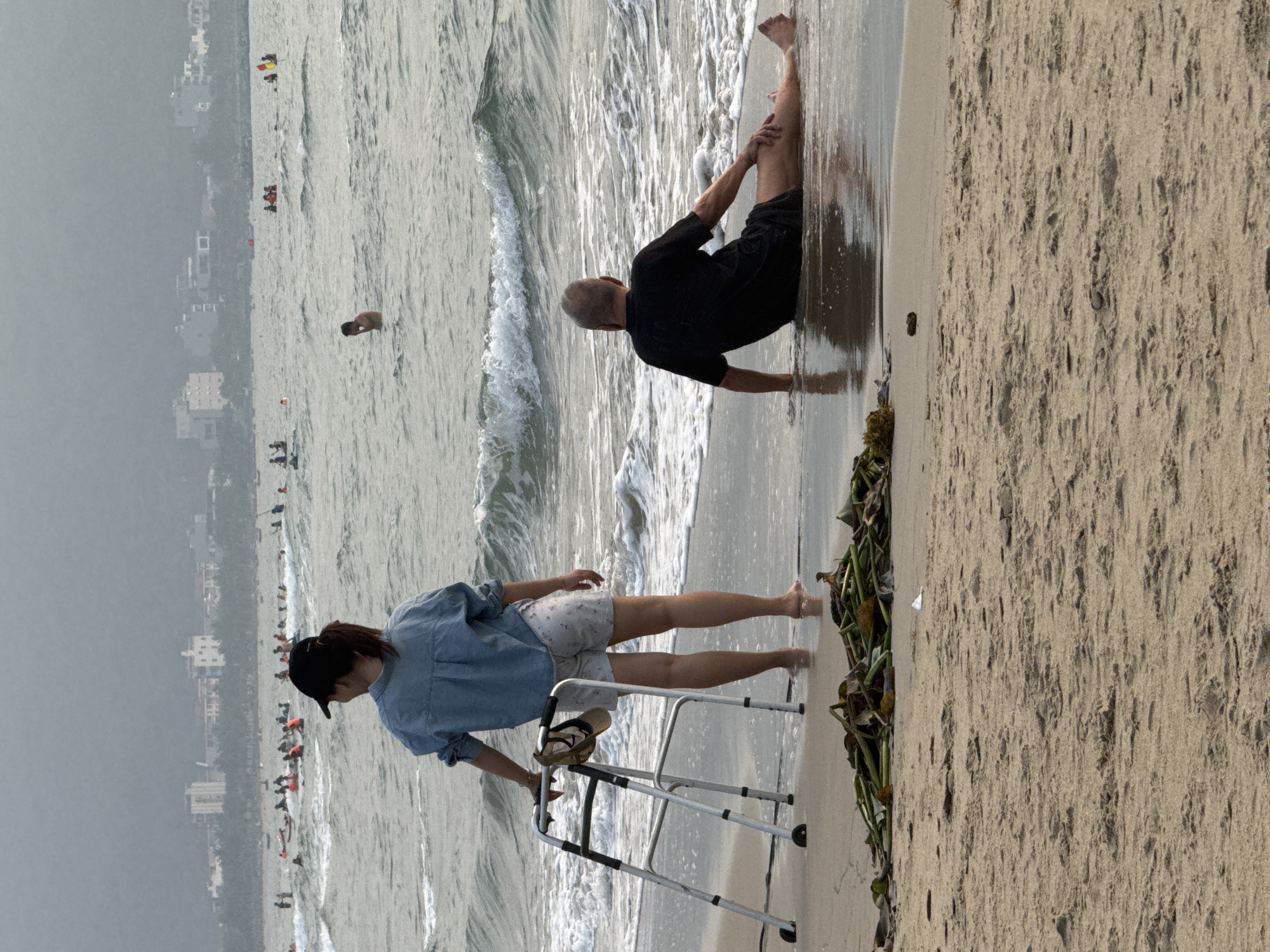An eye-level, medium shot photograph shows an overcast day at the beach in Đà Nẵng, Vietnam.
In the foreground, a woman in a short-sleeved blue shirt, patterned shorts, and a dark-colored hat is standing near the edge of the ocean. She is using a silver walker to help with balance. Sandals sit atop the walker. To the right of her, an older man in a black shirt and dark shorts is sitting in the water, seemingly stretching his leg.
The middle ground is filled with the ocean, which contains waves. Several people are swimming and standing in the water in the distance.
In the background, the city of Đà Nẵng is visible as a series of white buildings fading into the gray sky. The beach sand is tan and stretches along the right side of the frame. A line of seaweed is visible on the sand near the edge of the water.