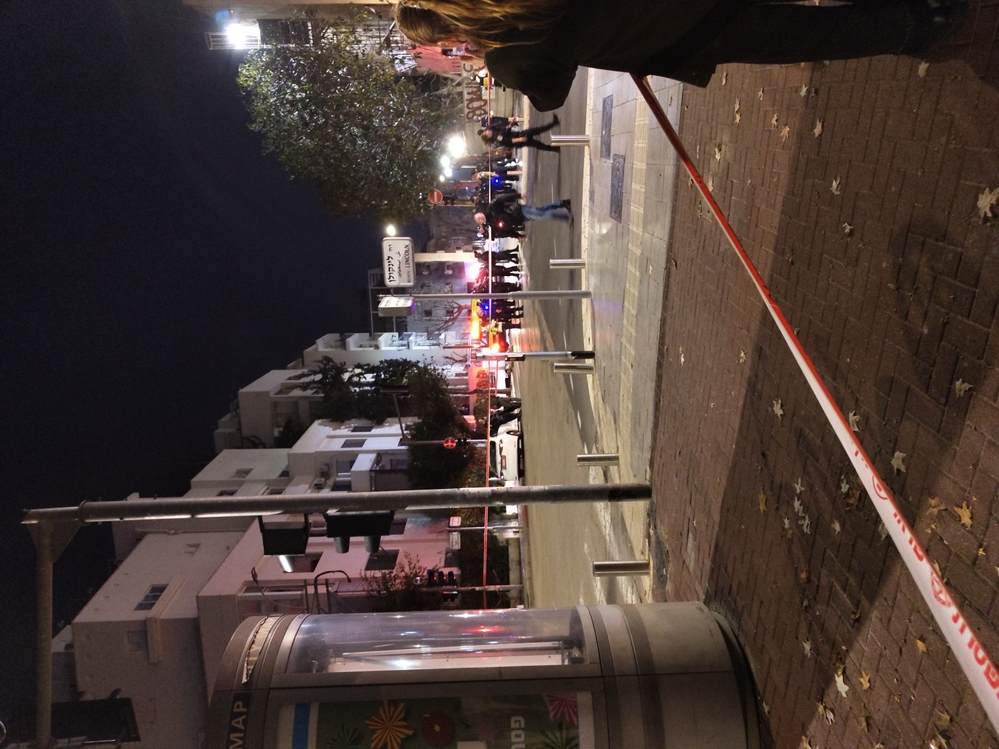 The image depicts a nighttime urban scene on Lincoln Street in Tel-Aviv, Israel, where an incident appears to have occurred.
In the immediate foreground, a red and white barrier tape, displaying Hebrew text including "משטרה" (Police), extends diagonally across a brick-paved sidewalk littered with scattered leaves. To the far left, a cylindrical street map kiosk is partially visible. The partial figure of a person with long, light brown hair stands in the lower right, observing the scene beyond the tape.
The midground reveals a street cordoned off, with multiple individuals, primarily in dark clothing, gathered around what appears to be an incident site. Flashing blue and red emergency vehicle lights are prominent in the center of the street, illuminating a group of people and at least one emergency vehicle. A white sedan is stopped or parked further down the street behind the main gathering. Street signs clearly indicate "רח' לינקולן" (Lincoln Street). A tall metal pole supports traffic lights, which appear to be red, and several silver bollards line the sidewalk.
In the background, multi-story buildings, predominantly white or light-colored, line both sides of the street under a dark night sky. Trees are visible between buildings and along the street, one large tree prominent on the right side. Streetlights provide general illumination to the area.
