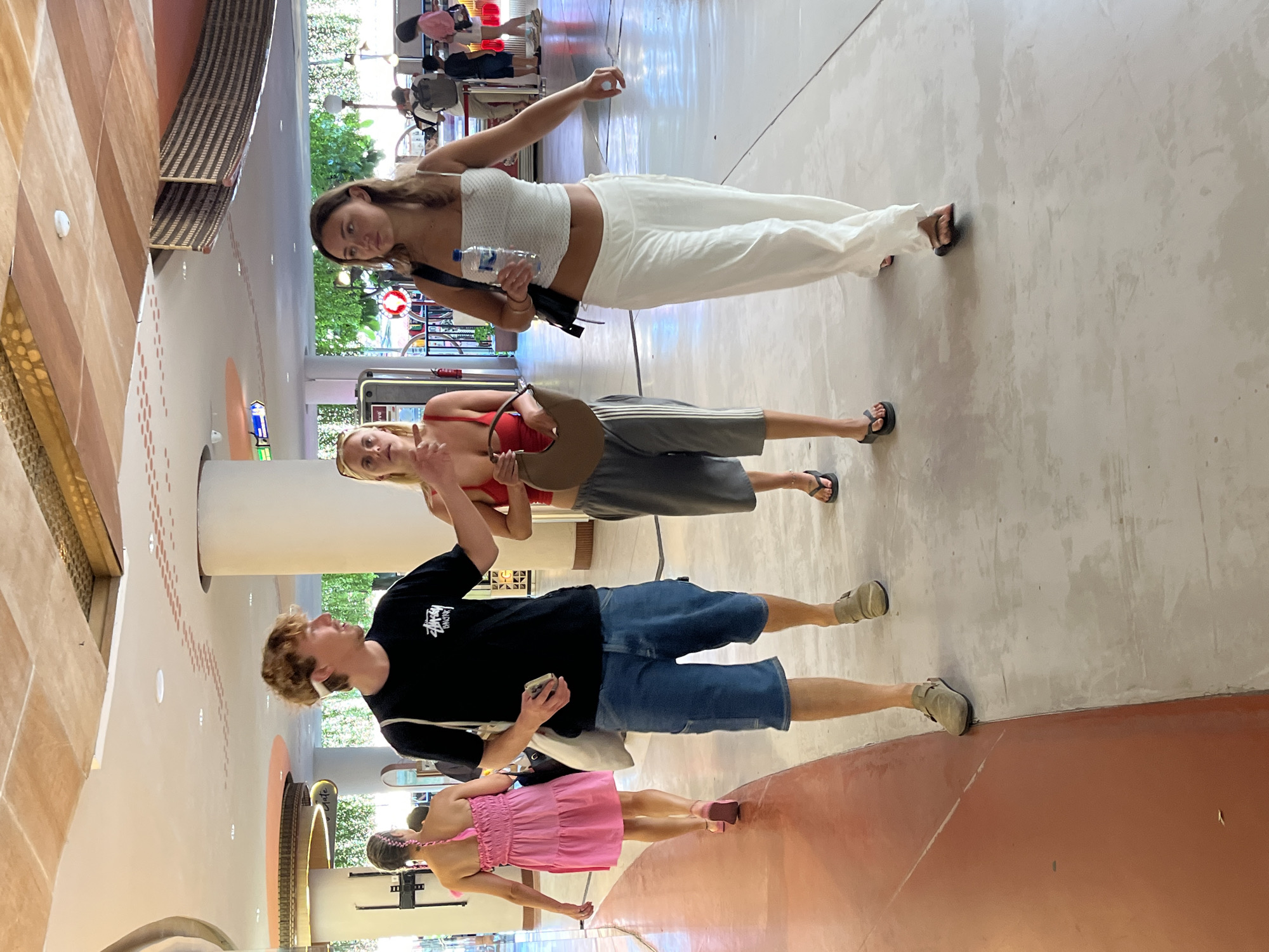 In Kuta, Indonesia, four individuals are observed walking in an indoor, open-air walkway with a textured ceiling featuring natural wood and cream-colored panels, possibly a commercial establishment or resort. On the left, a woman in a pink tiered dress with her hair in a braid walks away from the camera. In the center, a man wearing a black t-shirt and denim shorts walks toward the camera, holding a mobile phone. Beside him, a woman in a red bikini top and grey athletic shorts also walks toward the camera, carrying a brown purse. To the right, a woman in a white sleeveless crop top and loose white pants walks away from the camera, holding a plastic water bottle. Other pedestrians and architectural elements are visible in the background.