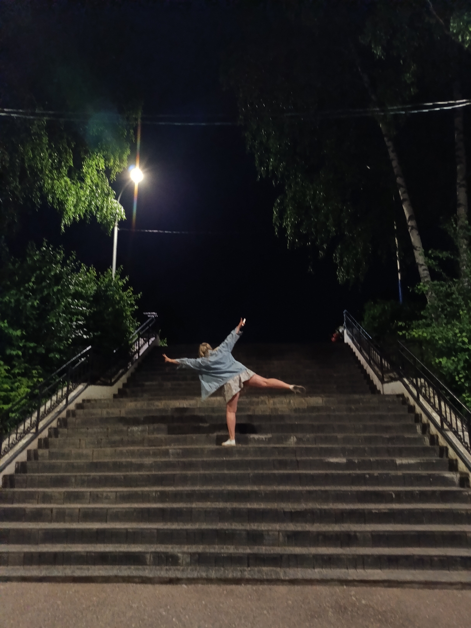 A person stands on a wide, outdoor staircase at night, facing away from the camera. They are balancing on their right leg with their left leg extended backwards and their arms outstretched horizontally. The person is wearing a light-colored denim jacket and a patterned skirt or dress, with white sneakers. The staircase is made of stone or concrete and has metal railings on either side. Trees with lush green foliage are visible on the left and right sides of the staircase, illuminated by a bright streetlamp positioned to the left of the frame. The background is dark, indicating nighttime. The scene appears to be located in a park or public area in the city of Chaikovsky, Russia, given the presence of well-maintained outdoor staircases and the type of vegetation.