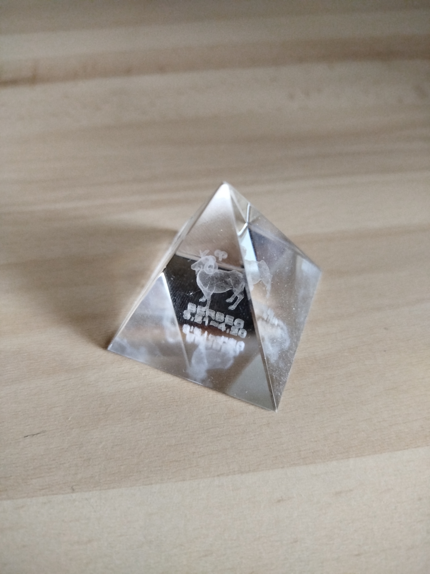 A clear, four-sided pyramid crystal is centered on a light brown wood-grain surface. The crystal has a laser-etched image of a reindeer and text. The text reads "RENDEER S.P.A.S.O. SWEDEN" in small block letters. The crystal is smudged and has imperfections on the surface. The location in Varna, Bulgaria is not evident from the image.