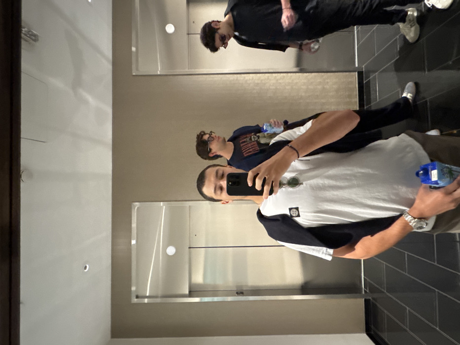 The image depicts three males in a modern interior hallway, likely a hotel or residential building lobby or common area, located in the city of Boston, United States. The scene is captured as a mirror selfie, with the primary subject holding a dark-colored smartphone to take the photograph.
The foreground subject is a male with short, dark hair, wearing a white t-shirt with a circular badge on the left sleeve (identifiable as Stone Island), a dark sweater draped over his shoulders, and sunglasses hanging from his collar. A silver wristwatch is visible on his left wrist, and he holds a blue, rectangular carton or bottle in his right hand.
Reflected in the mirror to the left of the foreground subject, slightly behind him, is a second male with dark, curly hair and eyeglasses, wearing a dark t-shirt with a graphic design and holding a clear plastic water bottle. To the right of the foreground subject, also reflected in the mirror and positioned further back, is a third male with dark, curly hair and sunglasses, dressed in a dark t-shirt and dark pants, holding a clear bottle.
The environment includes two stainless steel elevator doors on a light beige wall. The floor is composed of dark, rectangular tiles. Overhead, recessed lighting is visible in the white ceiling. The subjects appear to be waiting or passing through the area.