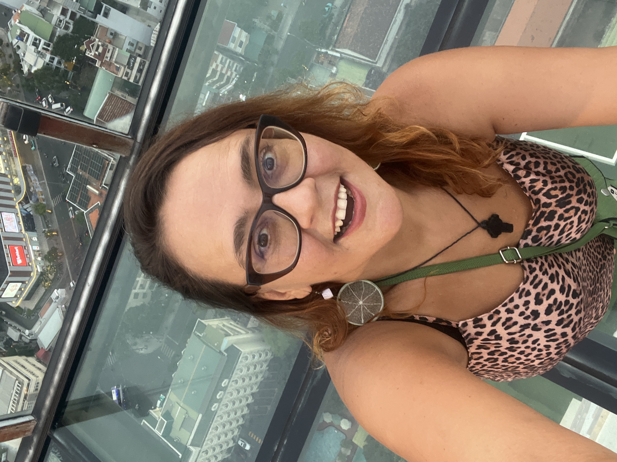 A woman with reddish-brown hair and glasses takes a selfie from a glass-floored observation deck overlooking the city of Nha Trang Ward, Vietnam. She is wearing a pink leopard-print camisole and a green crossbody bag. Her right ear sports a round earring that resembles a slice of lime. The glass floor reveals a cityscape with buildings, roads, and vehicles. The woman is smiling broadly, with her mouth open and teeth visible. The overall impression is one of excitement and enjoyment of the panoramic city view.