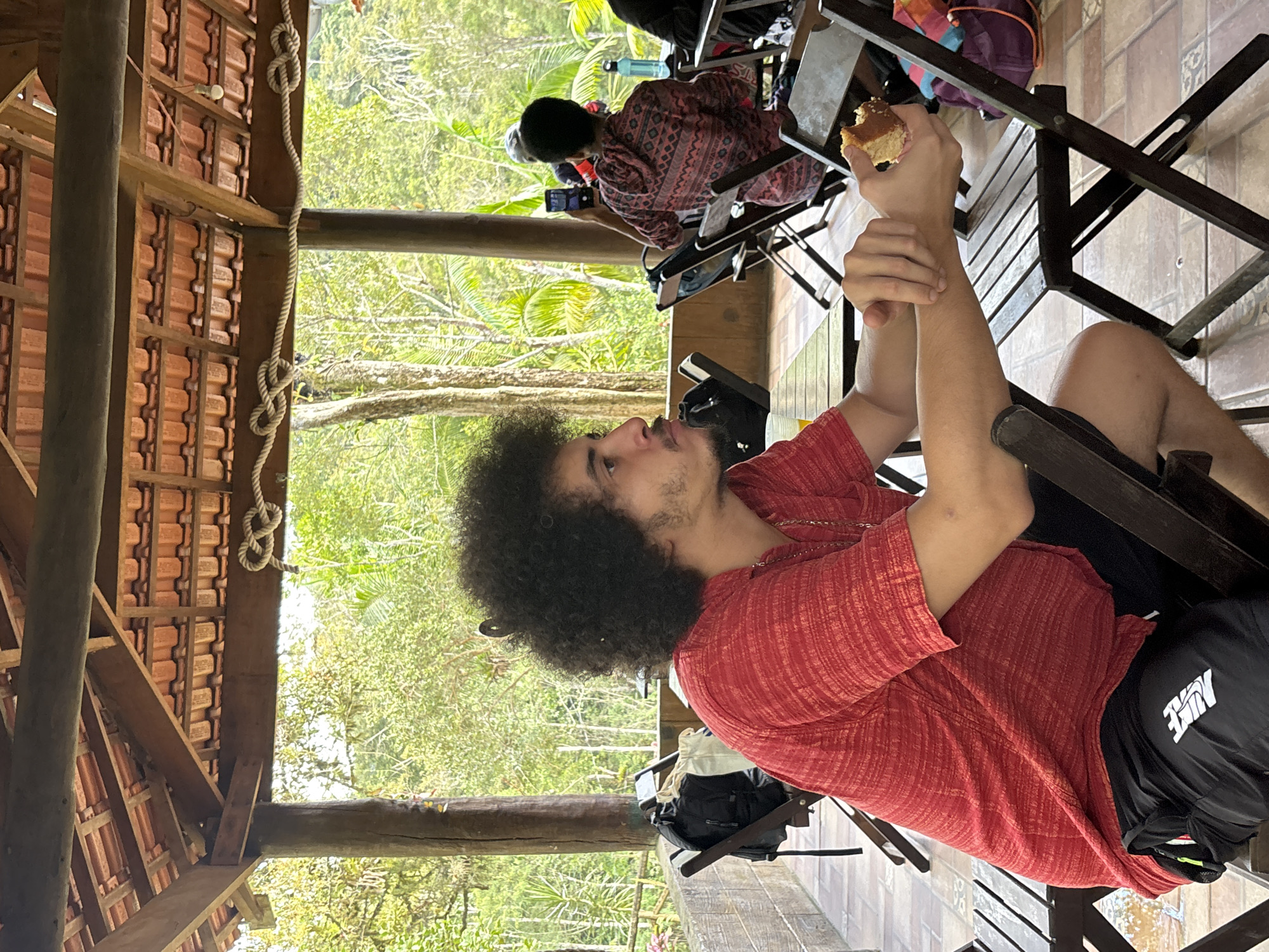 A person with dark, curly hair is sitting on a wooden chair, holding a piece of cake. They are wearing a red, short-sleeved shirt and black shorts. A black backpack with a white "Nike" logo is next to them on the chair. Other people are seated in the background, including one person in a patterned red shirt using a phone. The scene is outdoors, under a wooden structure with a tiled roof and a rope hanging from it. Trees are visible in the background. The location is in the city of São Paulo, Brazil.