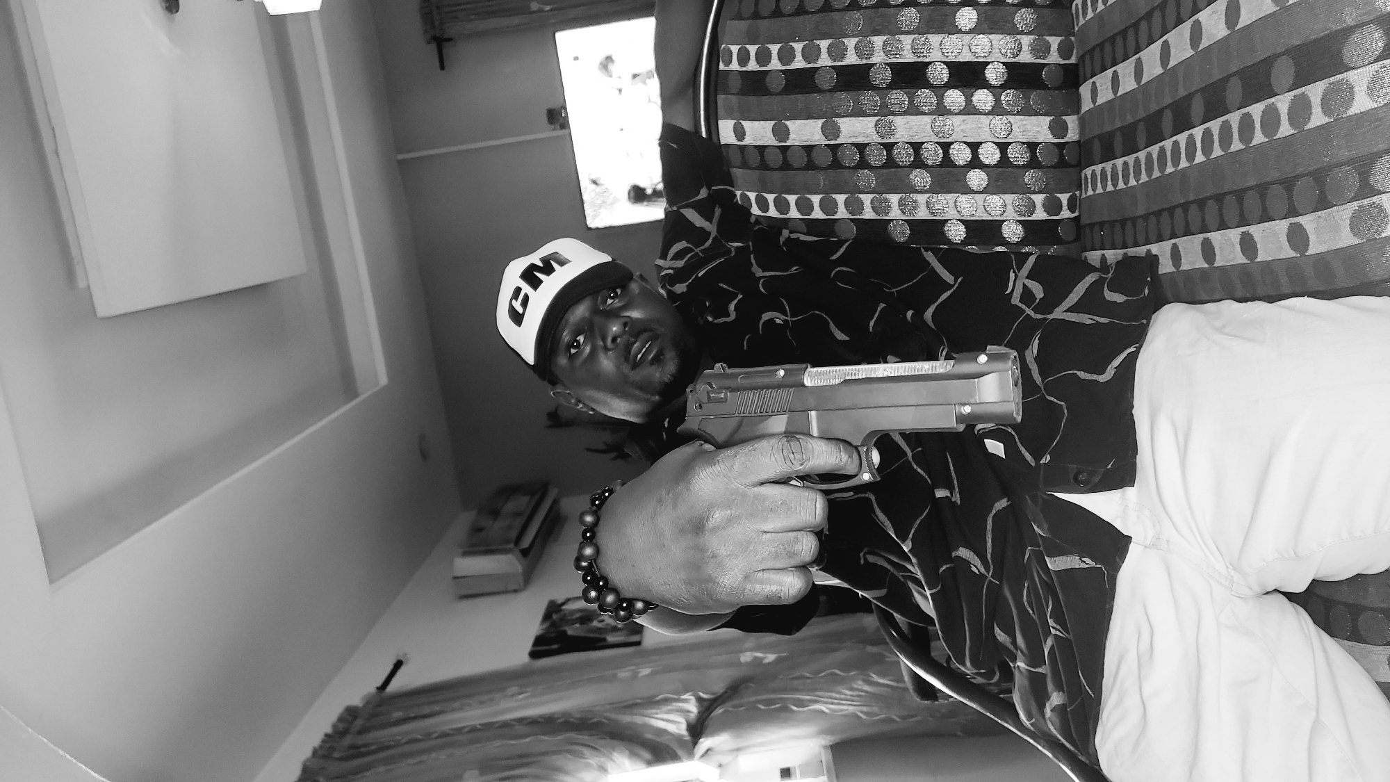 A black and white medium shot shows a Black man wearing a cap with "CM" on it, a patterned shirt, and white pants. He is seated on a couch and pointing a handgun forward towards the camera. The man has beads on his wrist. The background shows a living room with curtains, an air conditioning unit, and a television. The image is set in Narayi, Nigeria.