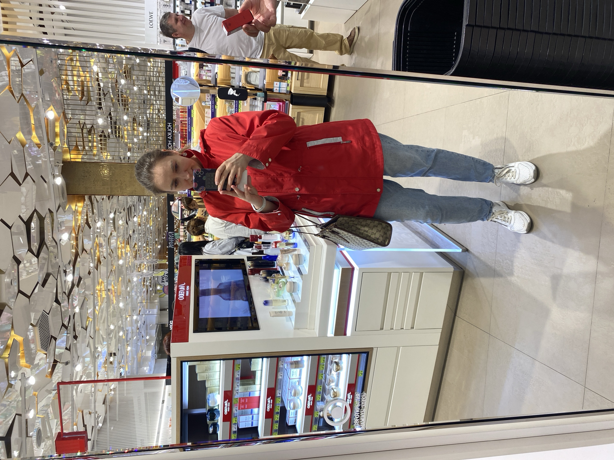 An image, presented sideways and mirrored, shows a woman taking a mirror selfie inside what appears to be a cosmetics or fragrance retail store. The woman, who is the primary subject, wears a red jacket, light blue jeans, and white sneakers, and holds a smartphone with a distinctive patterned case to capture her reflection. A patterned handbag hangs from her shoulder.
Behind her, also reflected in the mirror, stands a man in a white t-shirt and khaki pants, holding a small red box, seemingly browsing products. The store environment features bright artificial lighting, white display counters, and shelves showcasing various beauty products in bottles and jars. One display includes a digital screen. The store's interior design is modern, with walls and ceilings adorned with geometric, reflective panels, some featuring gold accents and embedded lights. The floor is covered with light-colored tiles. A large, black grated bin or display unit is visible to the right.
Visible text on product displays and signage, some of which is reversed in the mirror, includes "LOEWE" (a luxury brand), and several instances of "HNEIDO" (likely a brand name). Crucially, text in Russian, "НАЖАТЬ ЗДЕСЬ" (NAZHAT ZDES, meaning "CLICK HERE") and "ПЛЯР ОТКРЫТИЯ" (PLYAR OTKRYTIYA, likely part of "TESTER SAMPLE" or "OPENING SAMPLE"), confirms the location context of Yekaterinburg, Russia. The overall impression is a vibrant shopping experience within a high-end retail setting.