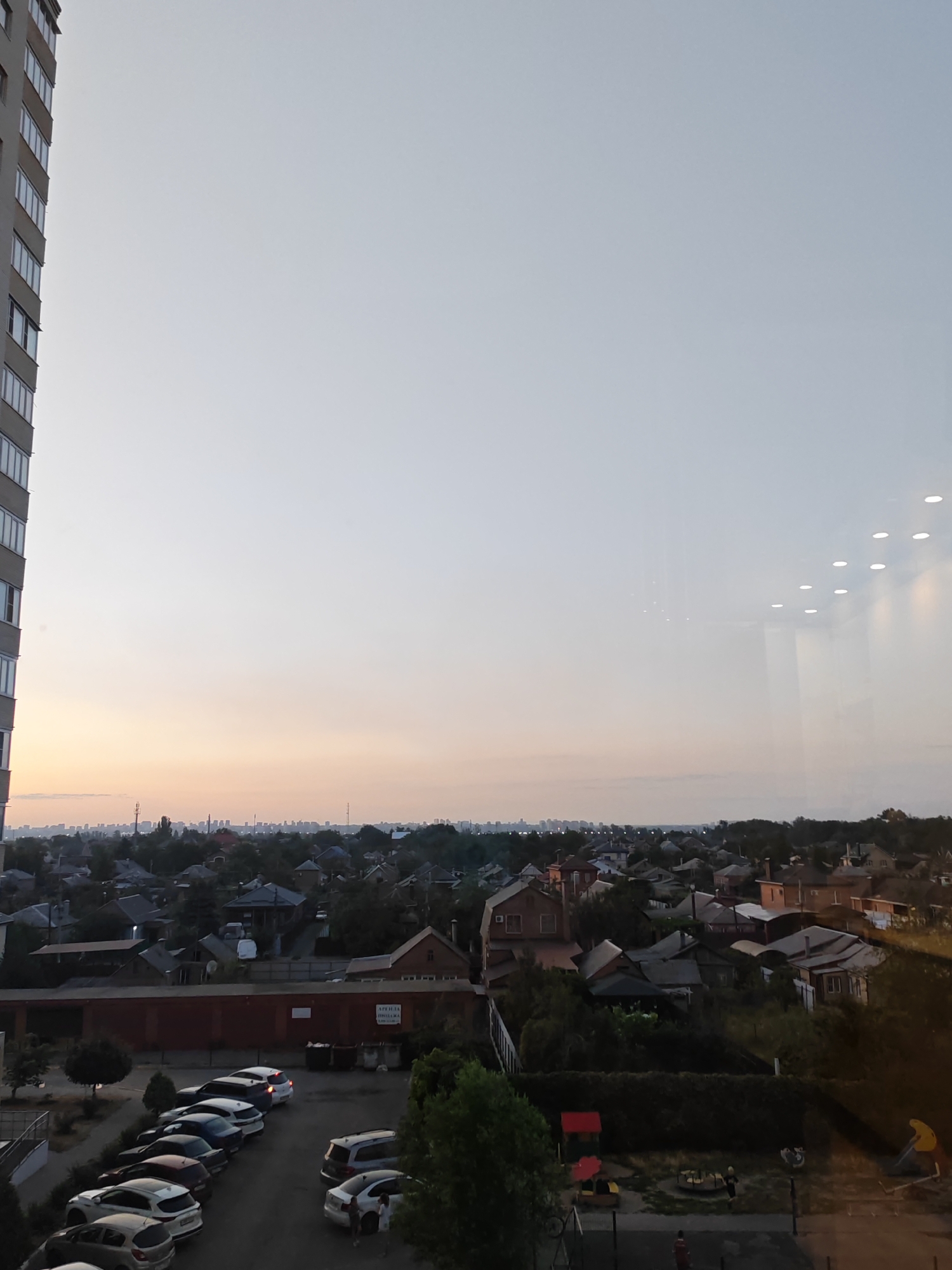 The image shows a view of the city of Bataysk, Russia, from a high vantage point, likely a building. In the foreground on the left is a tall, multi-story building with a light-colored facade and numerous windows. The sky occupies the upper half of the image, with a gradient of colors from a pale yellow near the horizon to a light gray at the top, suggesting it is either early morning or late evening.
Below the sky, a dense residential area is visible, characterized by numerous houses with varied rooflines, many of them appearing to be single-story or two-story structures. These houses are nestled amongst a significant amount of greenery, with trees and foliage interspersed throughout the neighborhood. A few commercial or industrial-looking buildings, with red brick facades and flat roofs, are also present in the middle ground. A parking lot filled with several cars is situated in the lower middle section of the image. A small playground area with a red play structure and a yellow slide is visible in the lower right portion. In the distance, a hazy cityscape can be seen on the horizon.