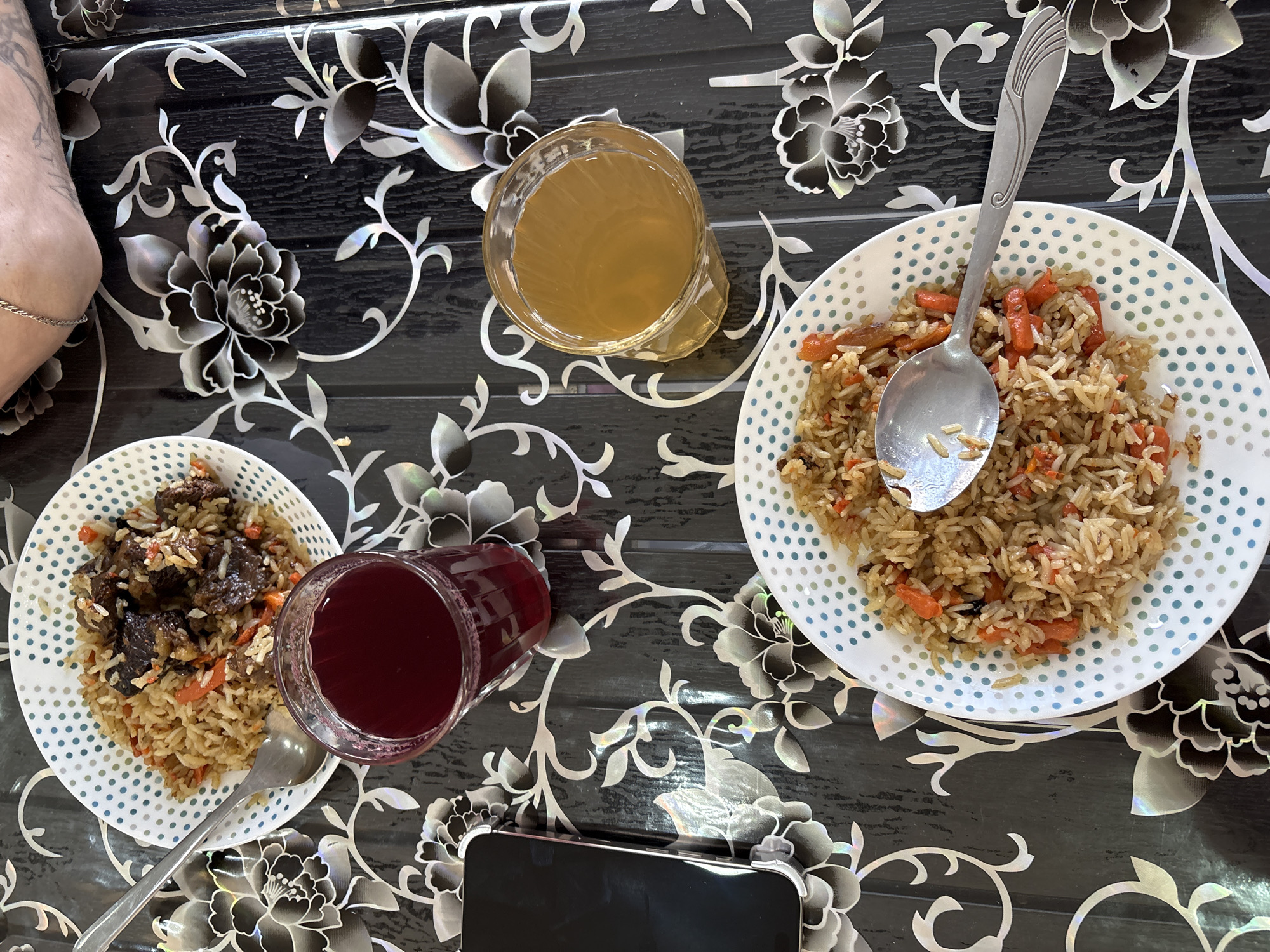 This image shows a table setting with two plates of rice pilaf, each with a spoon, along with two glasses of liquid, one clear and one red, and a smartphone. The pilaf on the left appears to contain meat, while the pilaf on the right appears to contain carrots. The table is black with a white floral pattern. The location is not evident from the image, and there is no indication it is in the city of Ust-Ilimsk, Russia. An arm is visible in the upper left corner of the photo.