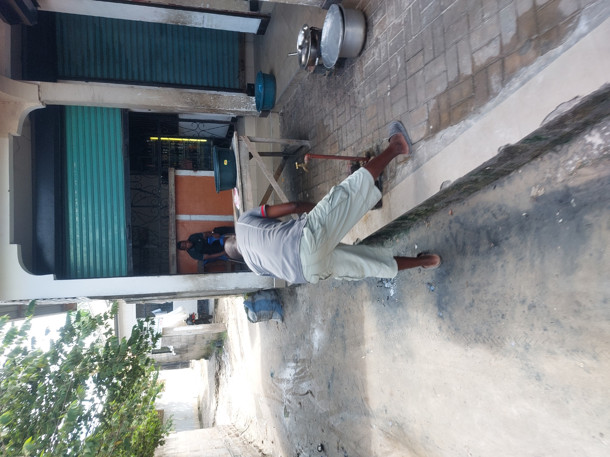 This outdoor image depicts a street scene, likely in a residential or commercial area of Dar es Salaam, Tanzania, with a mix of paved and dirt ground.
In the foreground, a man, viewed from behind, wears a light grey short-sleeved shirt with red and black trim, and light-colored shorts, along with sandals. He is positioned near a freestanding water tap with a red pipe, appearing to be using it or interacting with it. Several large metallic cooking pots, some lidded, are placed on the paved ground near the tap. A blue plastic basin sits further back on the paved area.
To the left, a building features two teal-colored roller shutters; one is closed, while the other is partially open, revealing an interior with a dark metal security gate and a hint of yellow light. Further back in the scene, another man is seated on a blue chair, wearing a dark shirt.
The ground transitions from a paved section with interlocking bricks, showing visible wet patches, to a dirt or sandy area further into the background, where faint tire tracks are discernible. Green foliage from trees is visible on the left side and in the distance. The overall lighting suggests it is daytime. No specific emotions are evident from the subjects, and there is no clearly legible text visible in the image.