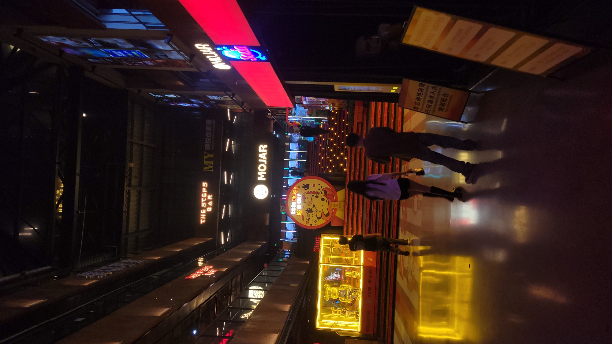 Here is a forensic description of the image:
The image captures a nighttime scene on a street in the city of Unknown, Unknown. Various neon signs illuminate the area. A red neon sign reading "MOJAR" is positioned at the top of the frame, with other signs for "MY" and "THE STEPS BAR" vertically arranged on the left side of the street. In the foreground, a large, round sign depicts a cartoon horse with Chinese characters above it. A storefront with a bright yellow neon display case is also present.
Three people are visible in the scene. Two of them, a man and a woman, appear to be walking side-by-side, while the third person is located further down the street and appears to be walking away from the camera. The street itself has a glossy surface, reflecting the surrounding lights and signs. The architecture suggests a modern commercial area. A sign near the front appears to have the following: "本店谢绝自带 任何酒水入内 谢谢配合"