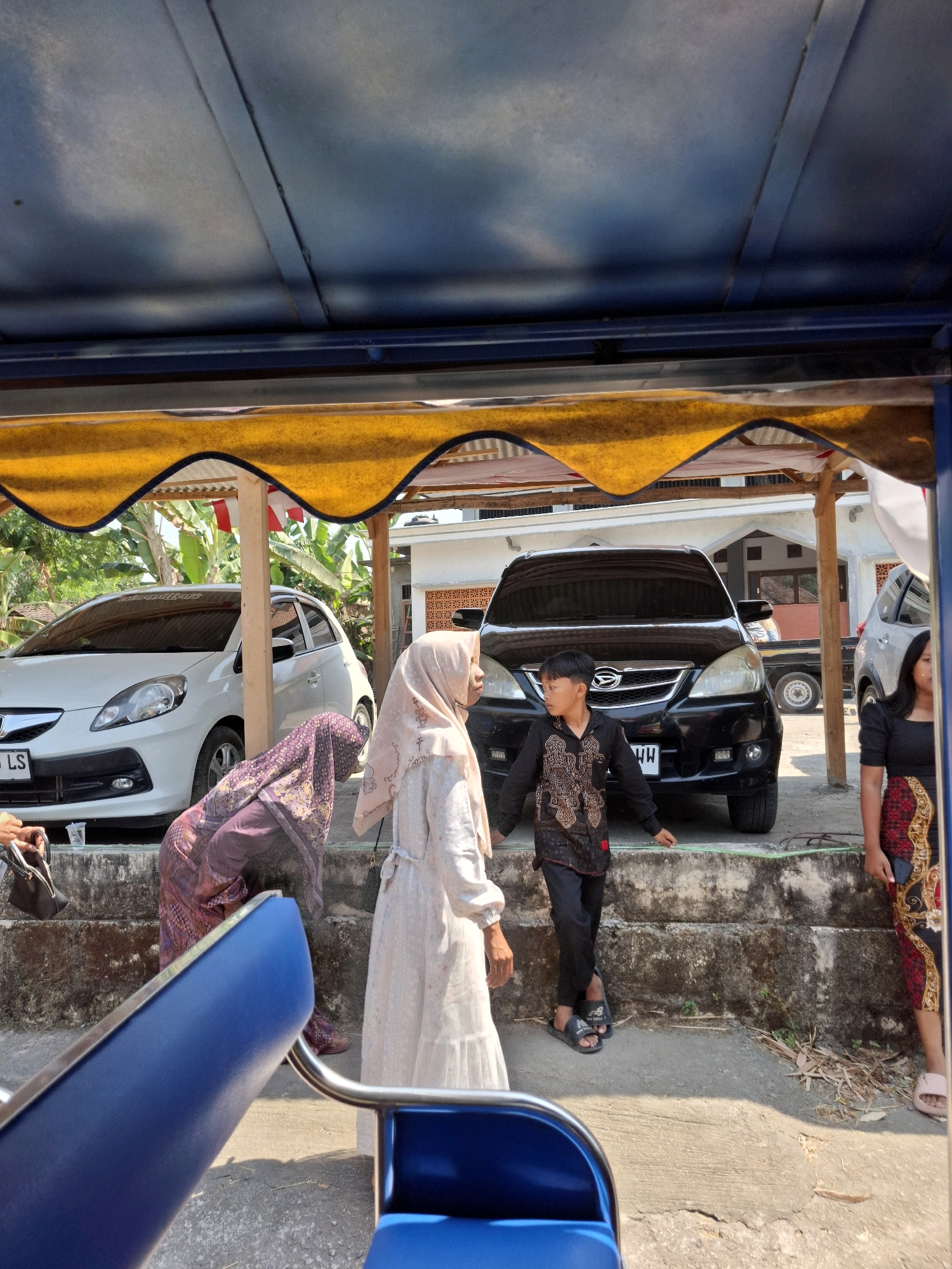 In Kedungmaron, Indonesia, a family is gathered outdoors under a yellow awning. The scene is viewed from inside a vehicle with blue seats, likely a mode of transport such as a pedicab or a tour vehicle. A white car and a black minivan are parked in the background.
Three women and one young boy are present. Two women, one in a purple and gold patterned hijab and dress, and another in a light-colored hijab and dress, are engaged in conversation near the white car. The third woman, wearing a black top and a patterned skirt, stands by the black minivan. The young boy, dressed in a patterned black shirt and black pants, stands facing the minivan and appears to be interacting with the woman in the black top.
The ground is concrete and dirt, with some dried leaves scattered around. The area appears to be a residential or parking area, with visible building structures and lush greenery in the background. The lighting suggests it is daytime, likely with bright sunlight.