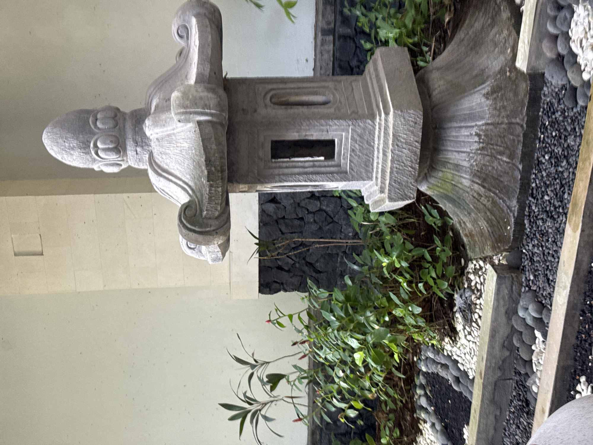 This is a close-up, eye-level, outdoor shot of a stone lantern in a garden setting in Kuta Selatan, Indonesia. The lantern is gray and appears to be made of carved stone. It has a tiered base with decorative carving, a rectangular middle section with an open window, and a decorative top with a finial. The lantern is positioned in the foreground, slightly off-center to the right.
Behind the lantern, there is lush green foliage with some small red buds or flowers. The plants are arranged in a garden bed that is bordered by stone and gravel. The gravel in the garden bed is a mix of dark gray and white pebbles. In the background, there is a wall constructed of light-colored stone blocks, with a section of dark, rough-hewn stone or rock directly behind the plants. The overall impression is one of a serene, landscaped garden, likely part of a resort or private residence in the tropical environment of Kuta Selatan. There are no people visible in the image.