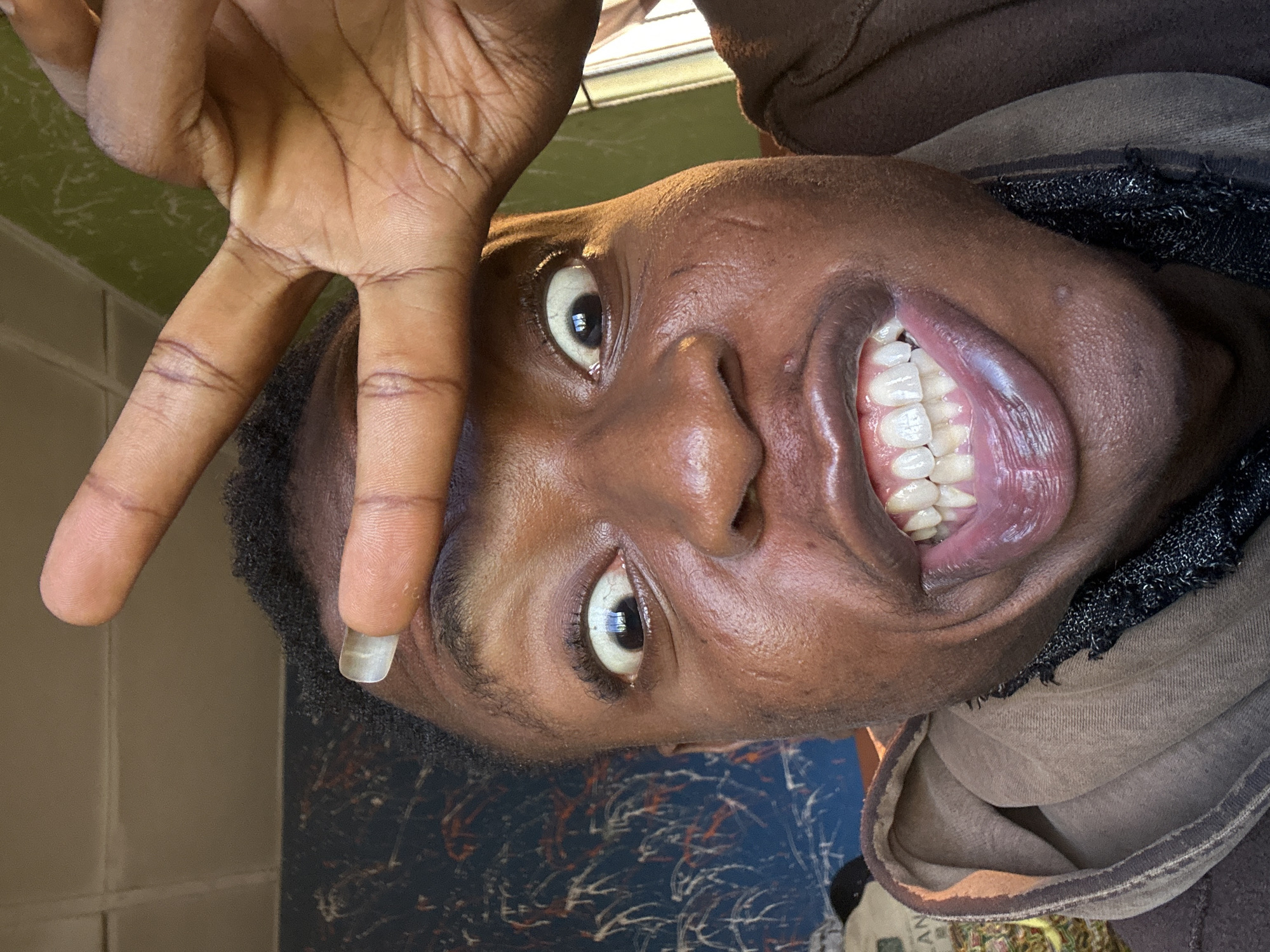 The image is a close-up, slightly tilted, selfie of a person with dark skin. They are making a peace sign with their left hand, held up near their forehead. Their eyes are wide open and their mouth is open, showing their teeth and tongue. They are wearing a brown hooded sweatshirt with a black woven neckline. The background is indistinct but appears to be a wall with a blue and white pattern, and possibly a glimpse of greenery. The location is Unknown, Unknown.