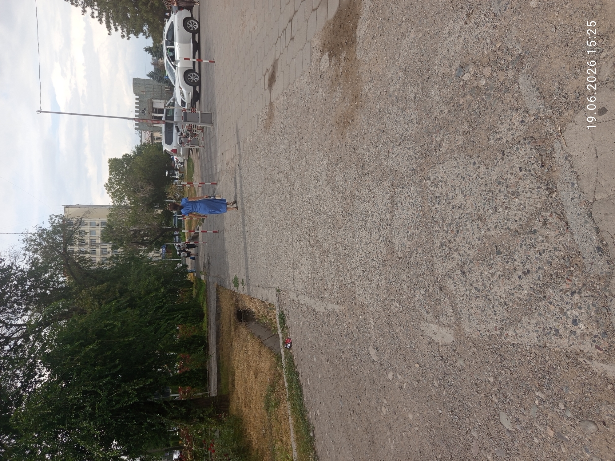 In Bishkek City, Kyrgyzstan, a woman in a blue dress walks away from the camera down a paved sidewalk. She carries a white handbag. The sidewalk is bordered by a gravelly area on the left and a paved street with parked cars on the right. The street has a white SUV and a white sedan. Trees line the left side of the sidewalk, and a tall building is visible in the background. The sky is cloudy. A boom barrier for a parking lot is also visible in the distance. The date stamp on the image reads "19.06.2026 15:25."