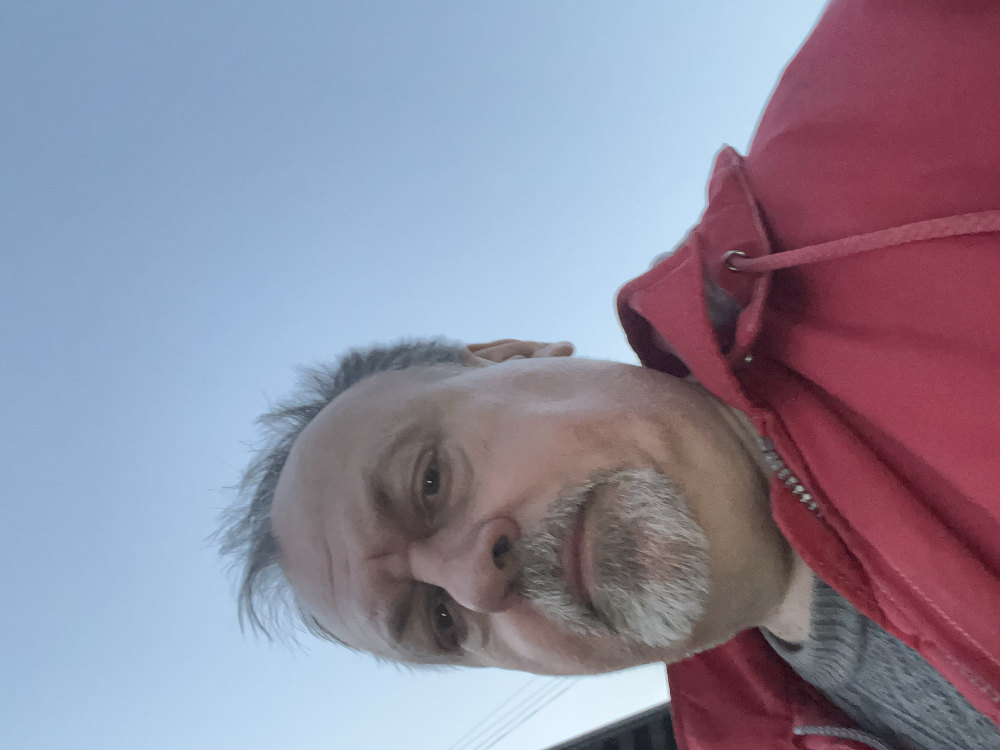 A middle-aged man with graying hair and a salt-and-pepper goatee is wearing a red jacket with a hood. The sky is visible in the background, suggesting an outdoor setting. The man is looking down towards the camera. The image is taken from a low angle. The location in Podolsk, Russia, is not discernable from the visual data.