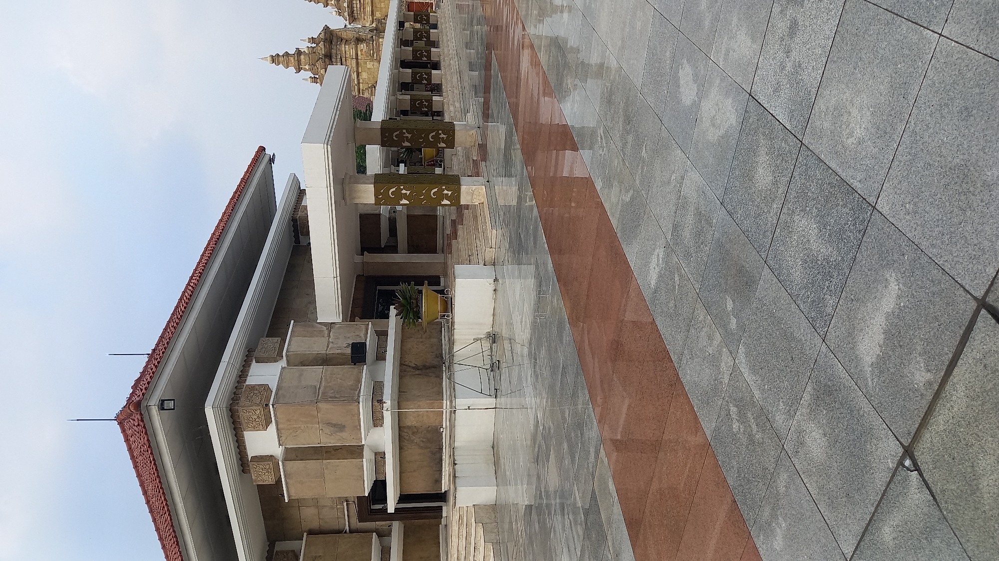 This is a photograph taken in the city of South Jakarta, Indonesia, depicting the exterior of the Taman Mini Indonesia Indah (TMII) cultural park. The image captures the facade of one of the park's pavilions, characterized by its tiered roof with red tiles and intricate stone detailing on the building's exterior. The pavilion features a prominent overhang supported by pillars decorated with a repeating pattern. In the foreground, a wet, tiled courtyard reflects the sky and the building, with a distinct band of reddish-brown tiles running diagonally across the paving stones. In the background, other structures with ornate spires, typical of Indonesian architecture, are visible, suggesting the cultural and diverse nature of the park. The sky is overcast, indicating a cloudy day. There are no people visible in the frame, and the scene appears calm and unoccupied. The lighting suggests it is daytime, likely early morning or late afternoon due to the diffused light.