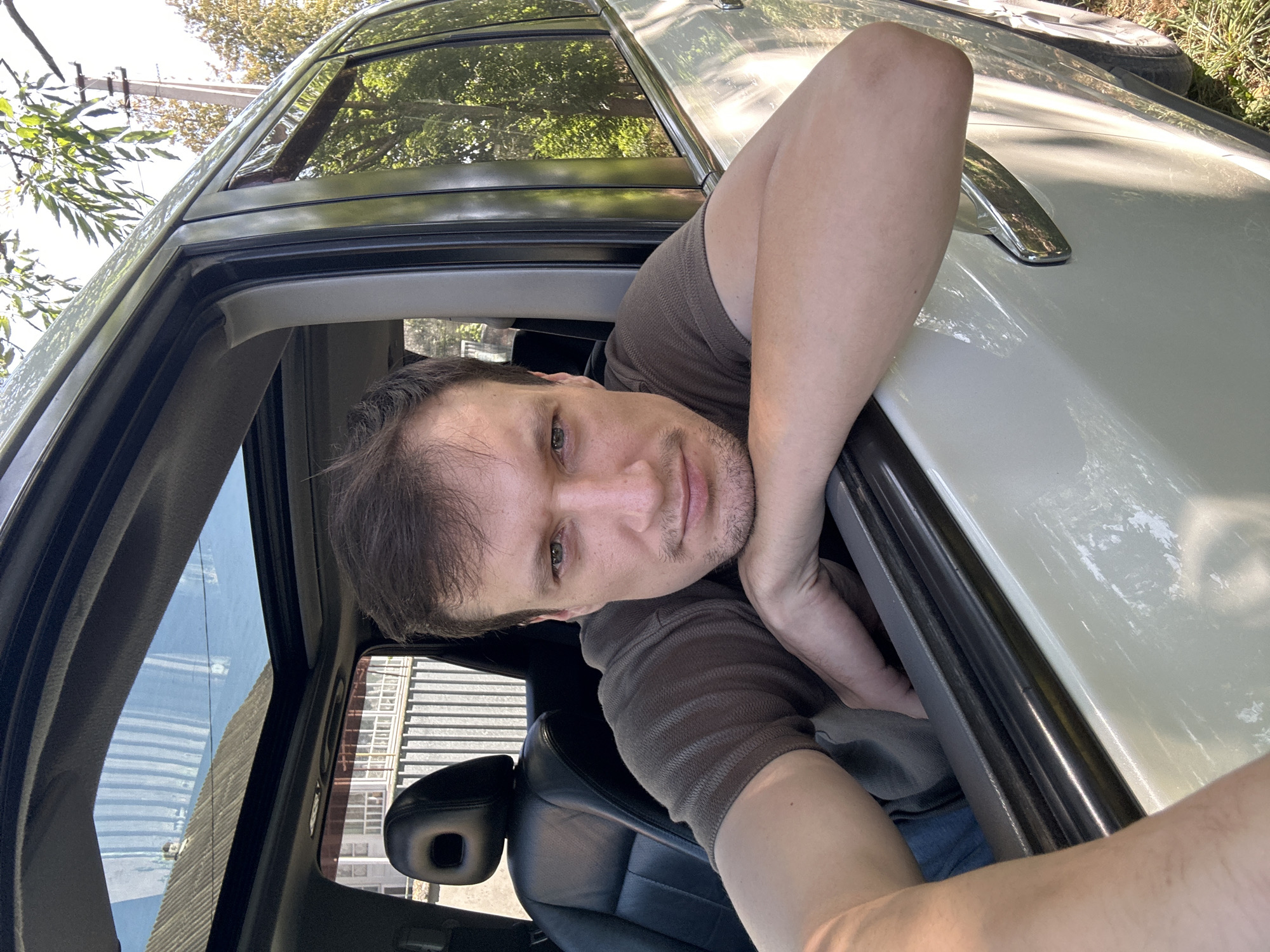 A medium-shot, over-the-shoulder selfie is taken from inside a vehicle, looking out the open driver's side window. The subject, a man with short brown hair and stubble, leans his forearms on the window frame, resting his chin on his hands. He is wearing a dark brown, short-sleeved, ribbed t-shirt. The car's interior features a black leather seat with a headrest. Outside the car, the background shows a fence, a residential building with a tiled roof, and trees with green foliage. The lighting suggests it is daytime, with shadows indicating a sunny environment. The location is identified as Makiv, Ukraine.
