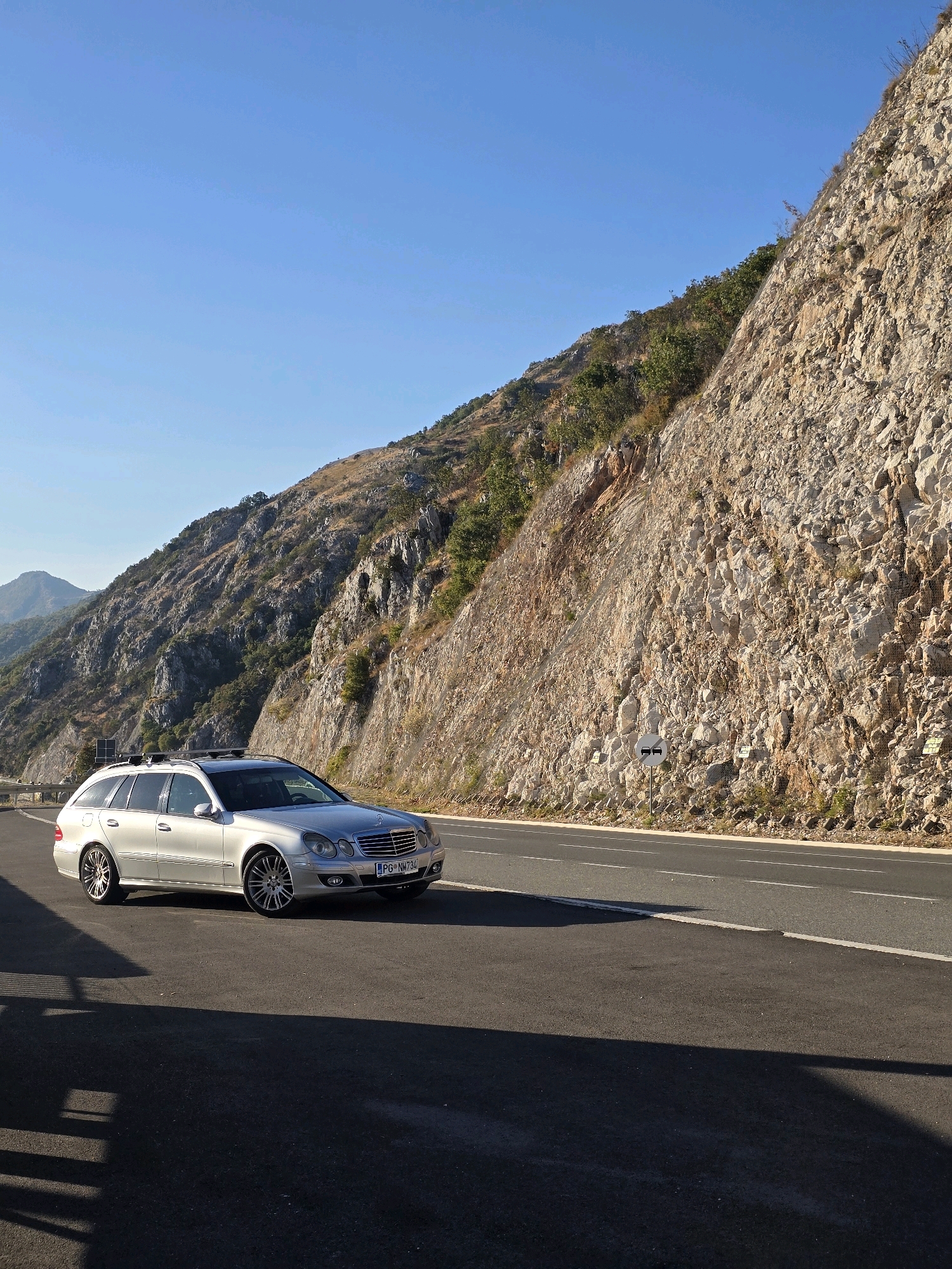 A silver Mercedes-Benz E-Class station wagon is parked on the side of a paved road that runs alongside a steep, rocky mountainside. The sky is clear and blue. The car is facing to the right of the frame and appears to be in good condition. The license plate reads "PG NM734". The road has clear white lane markings. The mountainside is covered in rough, tan-colored rock with sparse vegetation. There are some signs visible on the mountainside, including what appears to be a road sign with two cars on it. The sun is casting shadows on the road from the car and an unseen object to the left. The image appears to have been taken during the daytime in a mountainous, possibly rural or coastal area. This location is identified as Brajici, Montenegro.