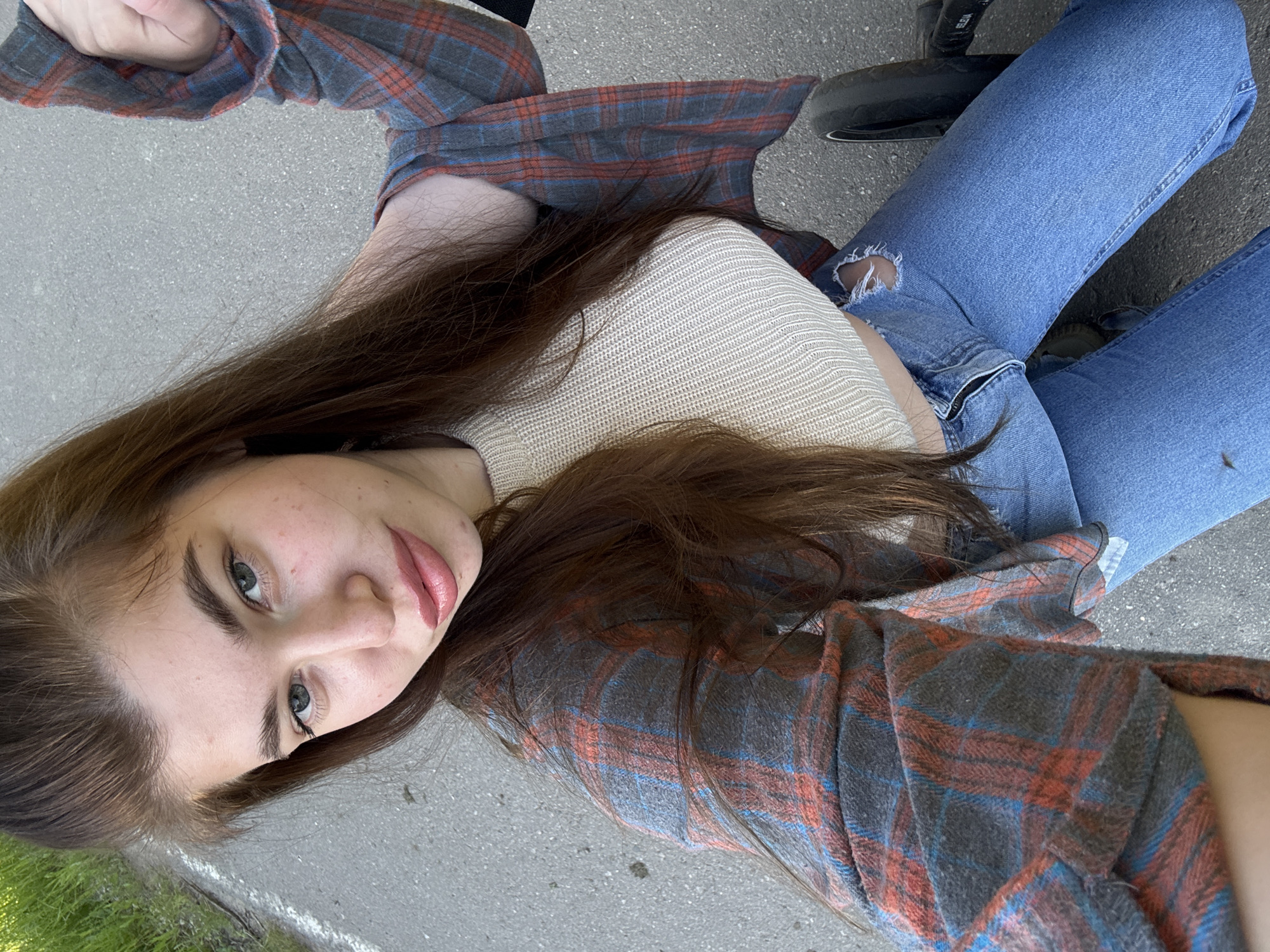 A young woman with long, dark brown hair and light-colored eyes is shown lying on an asphalt or concrete surface in an outdoor setting. She looks directly at the camera with a neutral expression, her lips adorned with pinkish-red lipstick, and light freckles visible on her cheeks and nose. She is dressed in a light, possibly cream or beige, knitted crop top, over which she wears an open plaid flannel shirt with a pattern of dark grey, red-orange, and blue. She also has on blue ripped jeans, with a noticeable tear on her left knee. In the background, glimpses of green grass and a white line on the ground suggest a paved area like a sidewalk or street. A dark object, which appears to be part of a scooter or bike, is visible near her right leg, bearing the text "SELMA". The scene is illuminated by bright, natural daylight, indicating it is daytime. The photograph captures the woman in a relaxed, reclined pose, taken from a slightly elevated perspective. The location context for this image is Мещёры, Russia.